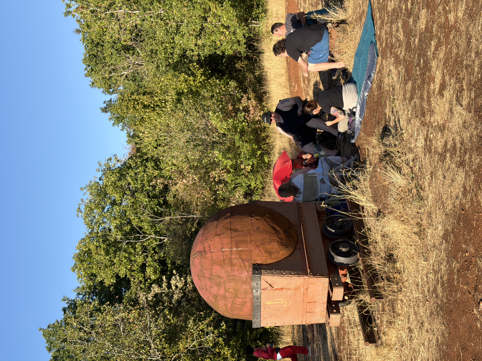 The image depicts an outdoor gathering in a rural or park-like setting. A group of individuals is seated on the ground and in chairs, some on blankets, in front of a large, rusty, spherical object mounted on a wheeled cart. The cart appears to be made of metal and has a weathered, industrial aesthetic, resembling a mine cart or similar equipment. The spherical object is reddish-brown and appears to be constructed from riveted metal panels. In the background, a dense line of trees with green foliage forms a backdrop, suggesting a wooded area. The ground is covered in dry, yellowish grass and reddish-brown soil, indicating a potentially arid environment or a dry season. The sky is clear and blue, suggesting fair weather. One person is visible in the distance on the left, walking away from the main group. The overall scene suggests a casual social gathering or a shared activity in a natural or historical industrial landscape. The location is identified as Schifflange, Luxembourg.
