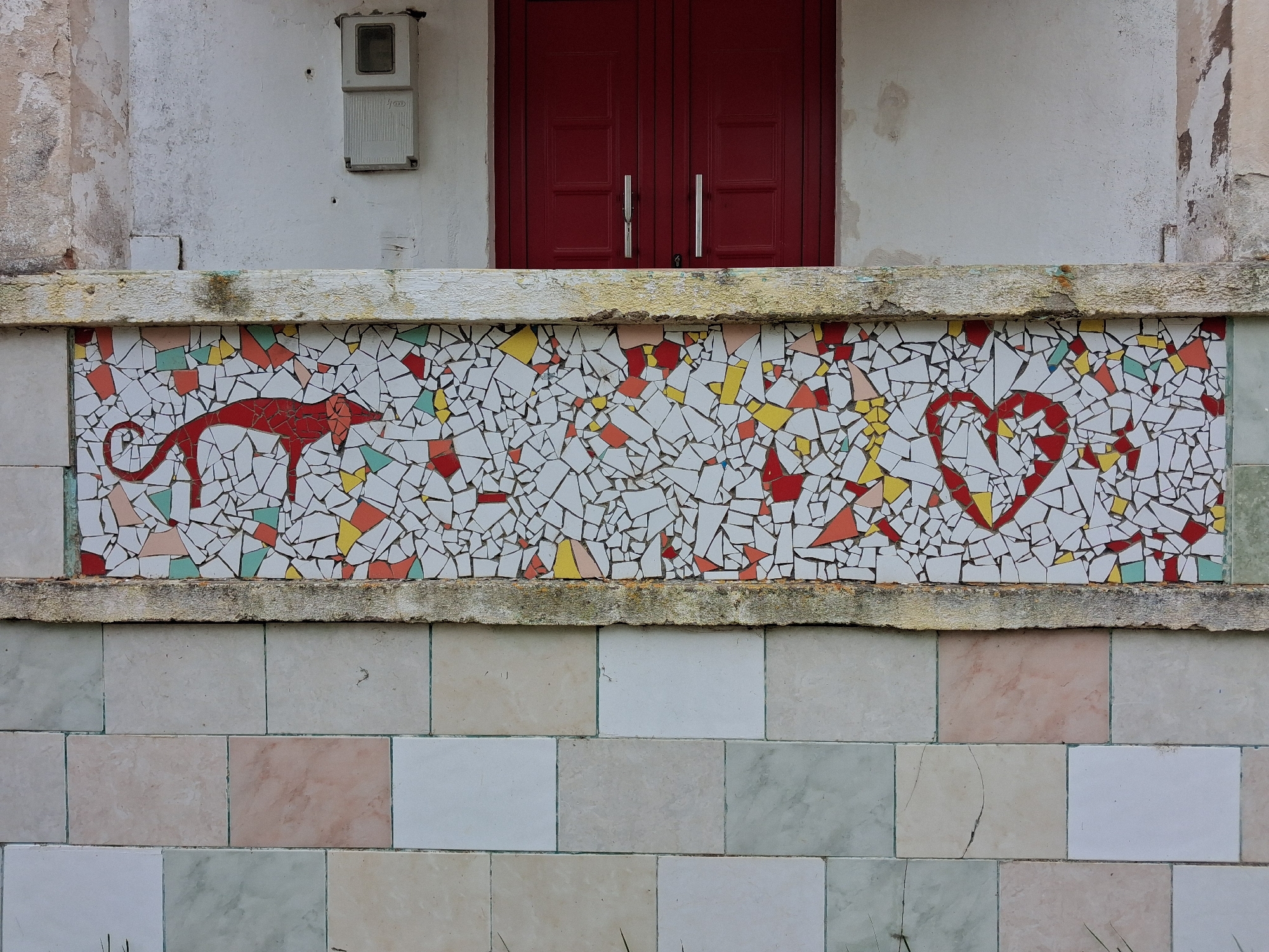The image shows a portion of a building facade. The upper part of the frame contains a white wall with peeling paint. At the top center of the image, there is a pair of closed, dark red doors with metal handles. Beneath this, a decorative tiled panel features a mosaic design. The mosaic depicts a red stylized dog or fox-like creature on the left, a scattering of red, yellow, and other colored tiles in the center, and a red heart shape on the right. The tiles appear to be broken and arranged in a pattern. Below the mosaic, a wall is covered in rectangular tiles of varying pastel colors. An electrical box is mounted on the wall above the mosaic and to the left of the doors. This image was taken in Soure, Portugal.