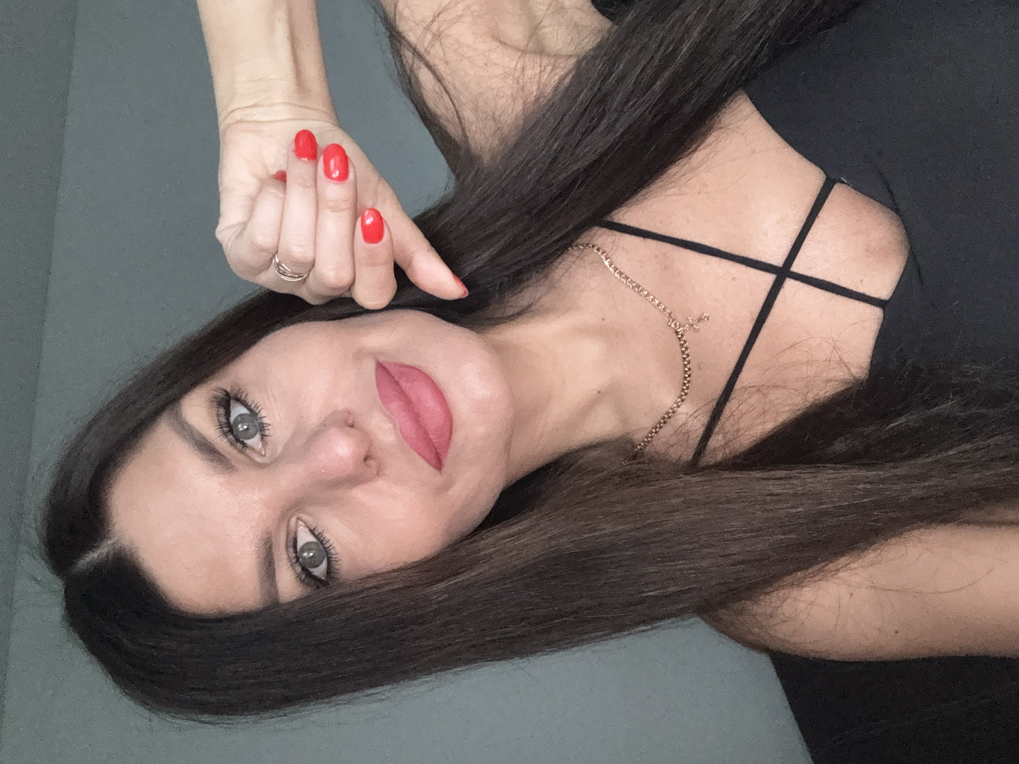 A woman with long dark hair and gray eyes is shown from the chest up, taking a selfie. She is wearing a black top with thin straps and a gold necklace with a cross pendant. Her nails are painted red, and she is wearing a ring on her left hand. Her left arm is bent at the elbow and her hand is held up near her face, with her thumb pointing downwards. The background appears to be an indoor setting with a plain gray wall. There is no indication of the location being in Chaikovsky, Russia.