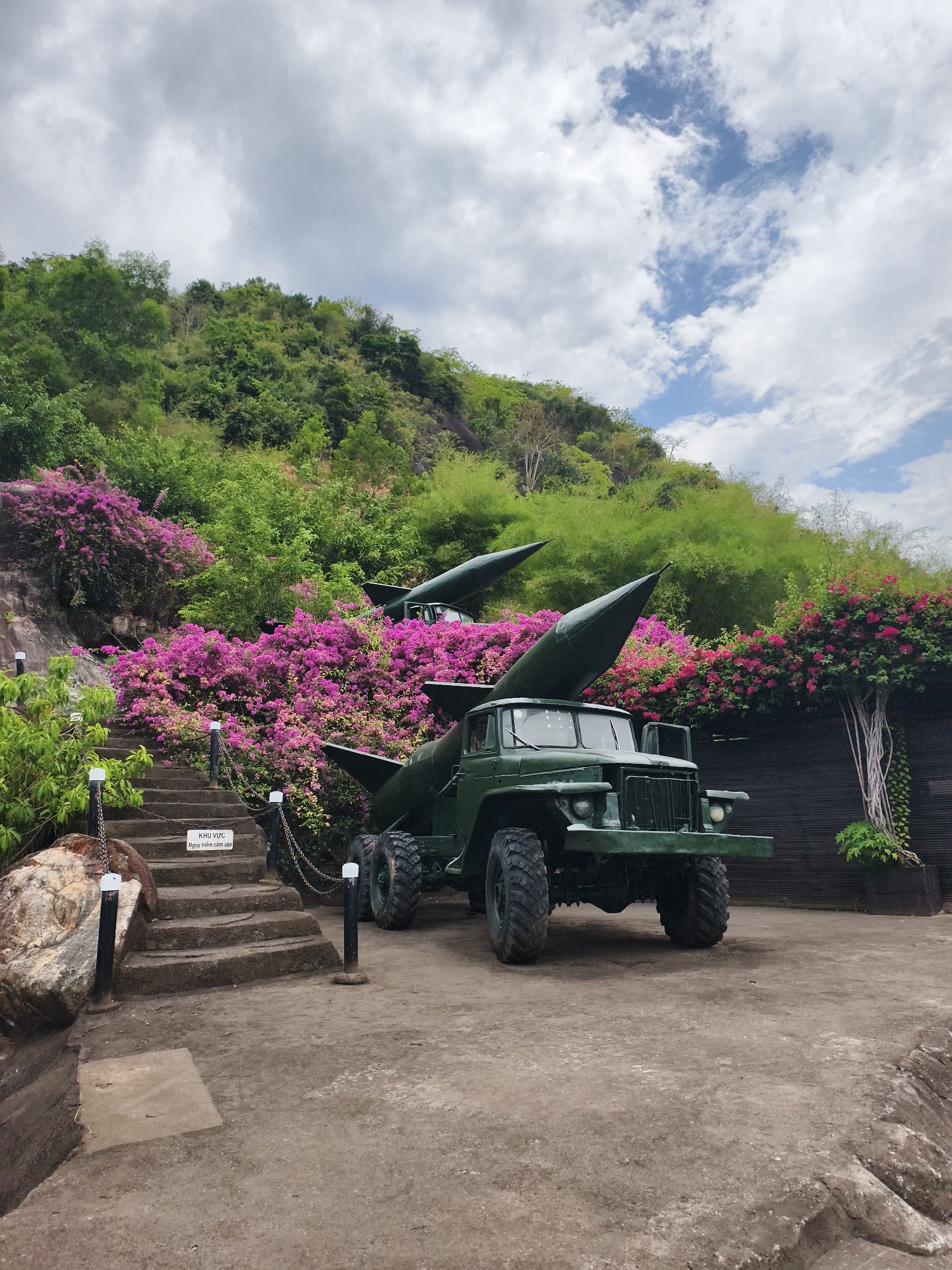 The image shows an outdoor display of a Soviet-era SA-2 surface-to-air missile launcher on a Zil-157 truck, located in Tay Nha Trang Ward, Vietnam. The missile carrier, painted in dark green, is parked on a dirt surface near a set of concrete steps leading up a hillside. The hillside is covered in lush green vegetation and vibrant pink bougainvillea bushes. In the background, the sky is filled with clouds. A wooden structure is to the right of the missile launcher. The sign is written in Vietnamese.