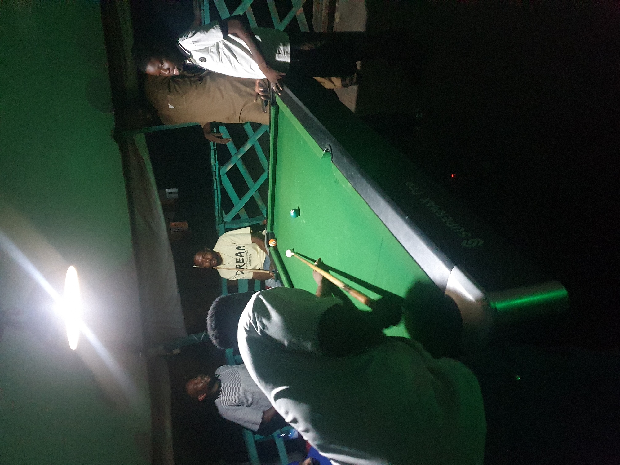 In Lokoja, Nigeria, a group of men are gathered around a pool table on a covered patio. The scene is dimly lit by a bright overhead lamp, casting long shadows. One man, wearing a light-colored shirt, is in the foreground, positioned to strike a white pool ball with a cue stick. He is focused on the game. To his right, a man in a yellow shirt with "DREAM" printed on it watches the game intently, holding a cue stick. Further back, two other men are visible. One, in a striped shirt, stands to the left, and another, in a light brown shirt, stands behind the man in yellow. The pool table is green, with a few balls on it. The background shows a railing and some indistinct structures, suggesting an outdoor or semi-outdoor entertainment area. The overall atmosphere appears casual and social, with the men engaged in a game of pool.
