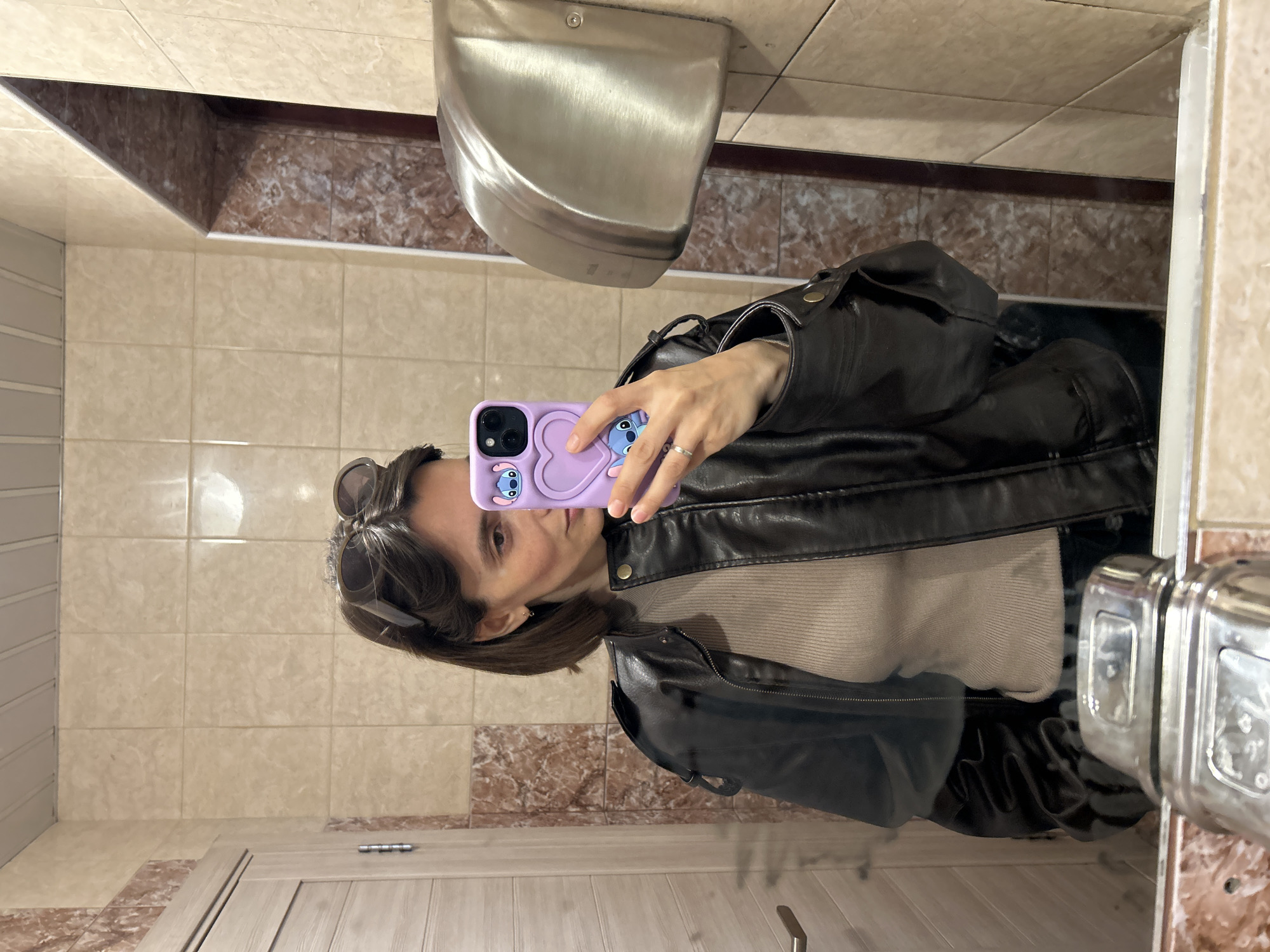 A person wearing a dark leather jacket takes a mirror selfie in a bathroom with light beige tiled walls and a narrow band of brown marble-like tiles along the top and bottom. A metal hand dryer is mounted on the wall above the person's head. The phone case is lavender with a cartoon character design. Sunglasses are perched on the person's head. The bathroom appears to be located in Moscow, Russia.