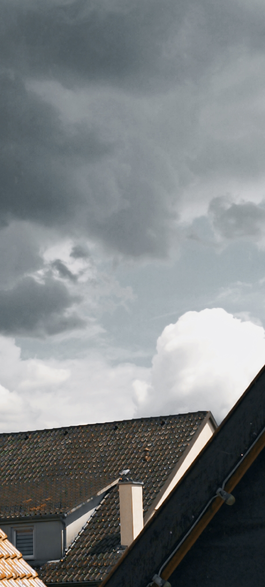 The image shows a rooftop view in Winnenden, Germany, with a dramatic sky overhead. Dark, heavy clouds dominate the upper portion of the frame, with lighter, cumulonimbus clouds visible towards the right. In the foreground, the tiled roofs of houses are visible, one featuring a light-colored chimney. A window with horizontal blinds is visible on the left side of one of the buildings. The perspective is from an elevated position, looking down at the rooftops and up at the sky. The lighting suggests an overcast day, possibly before or during a storm.