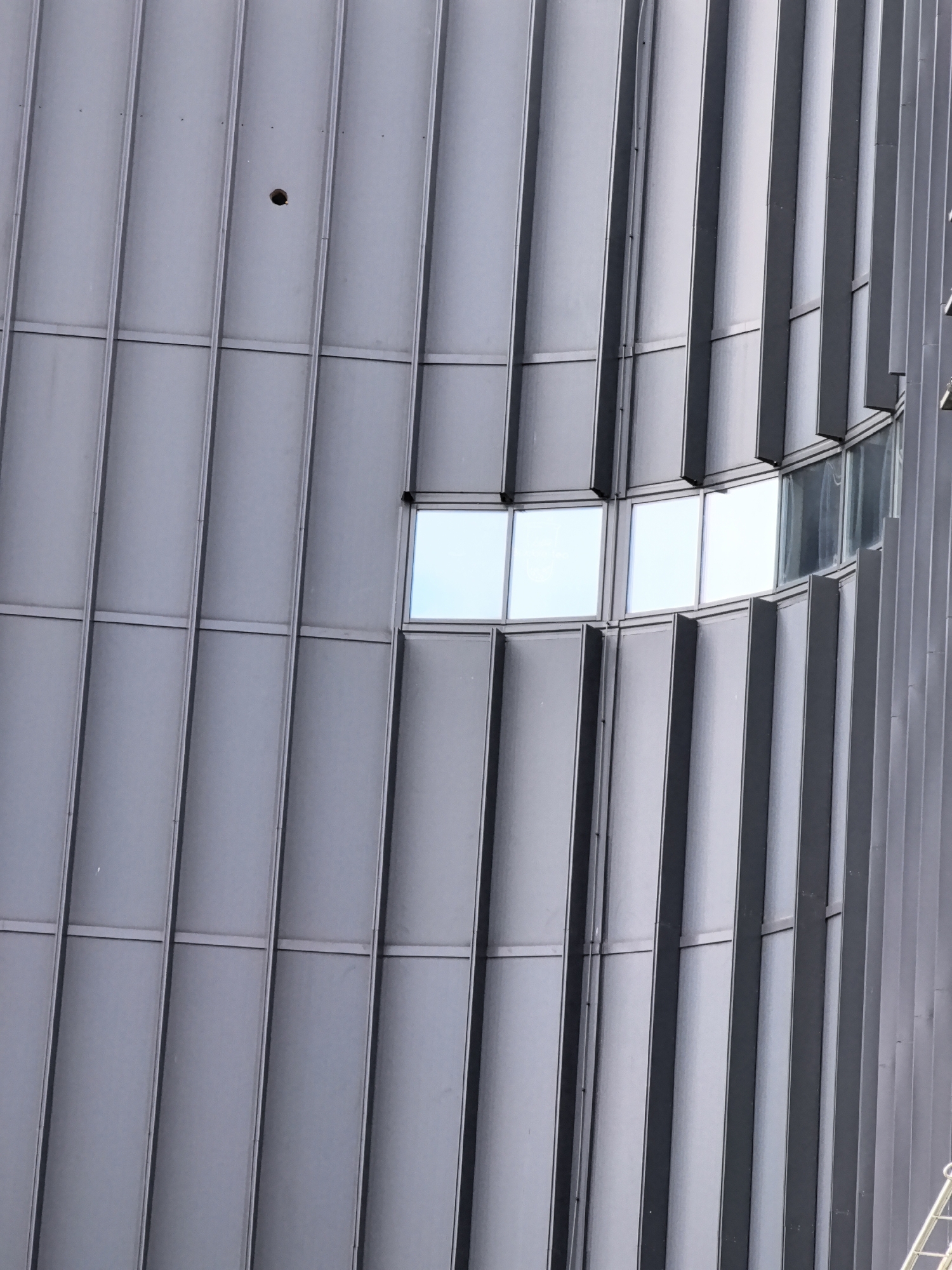 This media file is a close-up, eye-level, vertical shot of the exterior of a modern building. The building's facade is characterized by a repetitive pattern of vertical metallic panels, creating a ribbed effect. The color palette is predominantly gray and muted blue, suggesting an overcast or diffused lighting condition. The time of day is indeterminate due to the lack of strong shadows.
The most prominent features are the metallic panels and a small section of windows. The windows are rectangular and reflect a bright, possibly sky-blue light. One window, located centrally and slightly to the right of the frame, appears to be made of multiple panes, with some showing a slight curve in their arrangement. The metallic panels are uniformly colored and show minimal texture, emphasizing their smooth, manufactured quality. There is a single, small, dark circular hole visible in one of the metallic panels near the top left of the image.
The overall impression is one of architectural design and structural form. There are no people or identifiable objects other than the building itself. The scene is static and architectural, with no specific activity or event taking place. The location is not identifiable through any landmarks or distinctive features, making it a generic representation of a modern urban structure. The lack of human presence and the minimalist aesthetic create a sense of detachment or perhaps urban anonymity. No text is visible in the image.