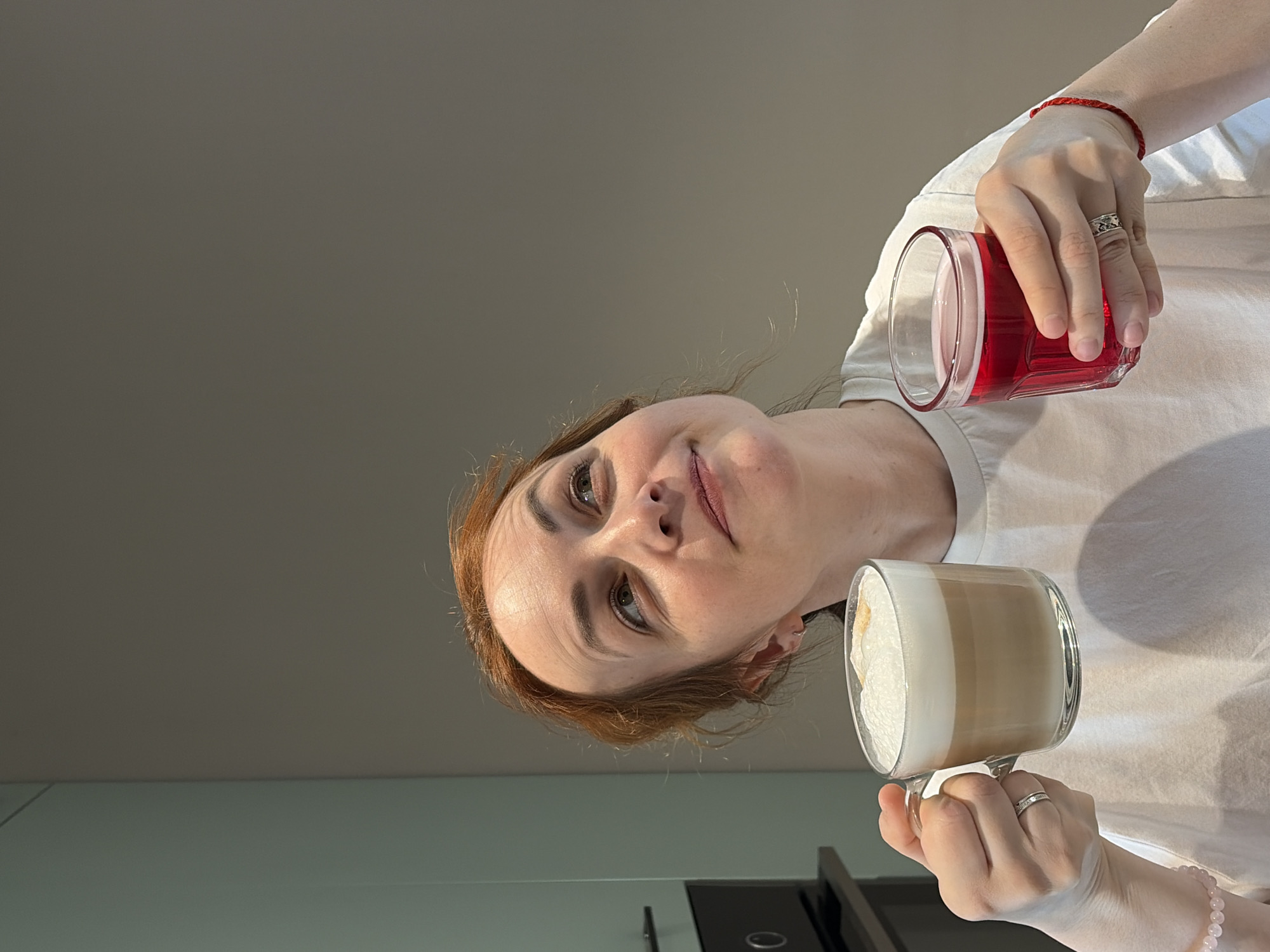 A woman with auburn hair is holding two glasses, one with a red liquid and the other containing a latte. She wears a white t-shirt and rings on her fingers. Her location is indeterminable from the given image. There is no evidence to suggest she is in Chaikovsky, Russia. The background appears to be a wall, and a countertop or appliance is visible near the bottom edge of the image. The image is taken from a low angle looking upward.