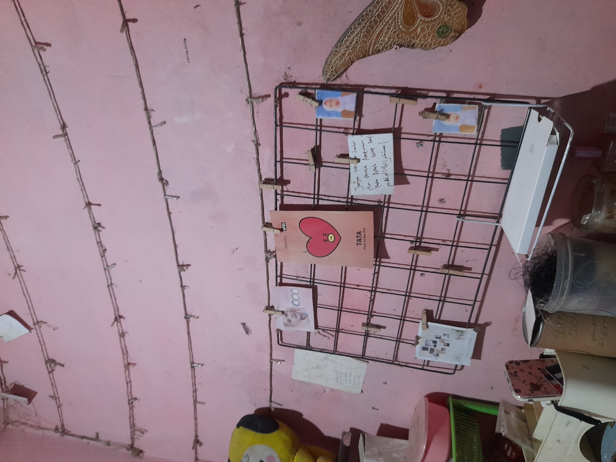 This image shows a wall decorated with a grid of photos and a string of fairy lights. The wall is painted a light pink color, and a large, decorative object with a patterned texture is hanging on the upper right side. The grid, made of black metal wire, is holding several small photos and a handwritten note. One of the photos shows a person wearing a headscarf. Another is a graphic of a red heart with the word "TATA" on it. A small, white card with multiple tiny photos is also attached to the grid. The fairy lights are draped vertically down the wall in several strands, casting a warm glow. To the right of the grid, there are some storage containers and other items, including a roll of something that appears to be paper or fabric. In the lower left corner, a yellow stuffed toy with black accents is partially visible. The scene appears to be indoors, likely in a bedroom or personal space. The lighting suggests it might be evening or night. The handwritten note is in Indonesian and Arabic script.