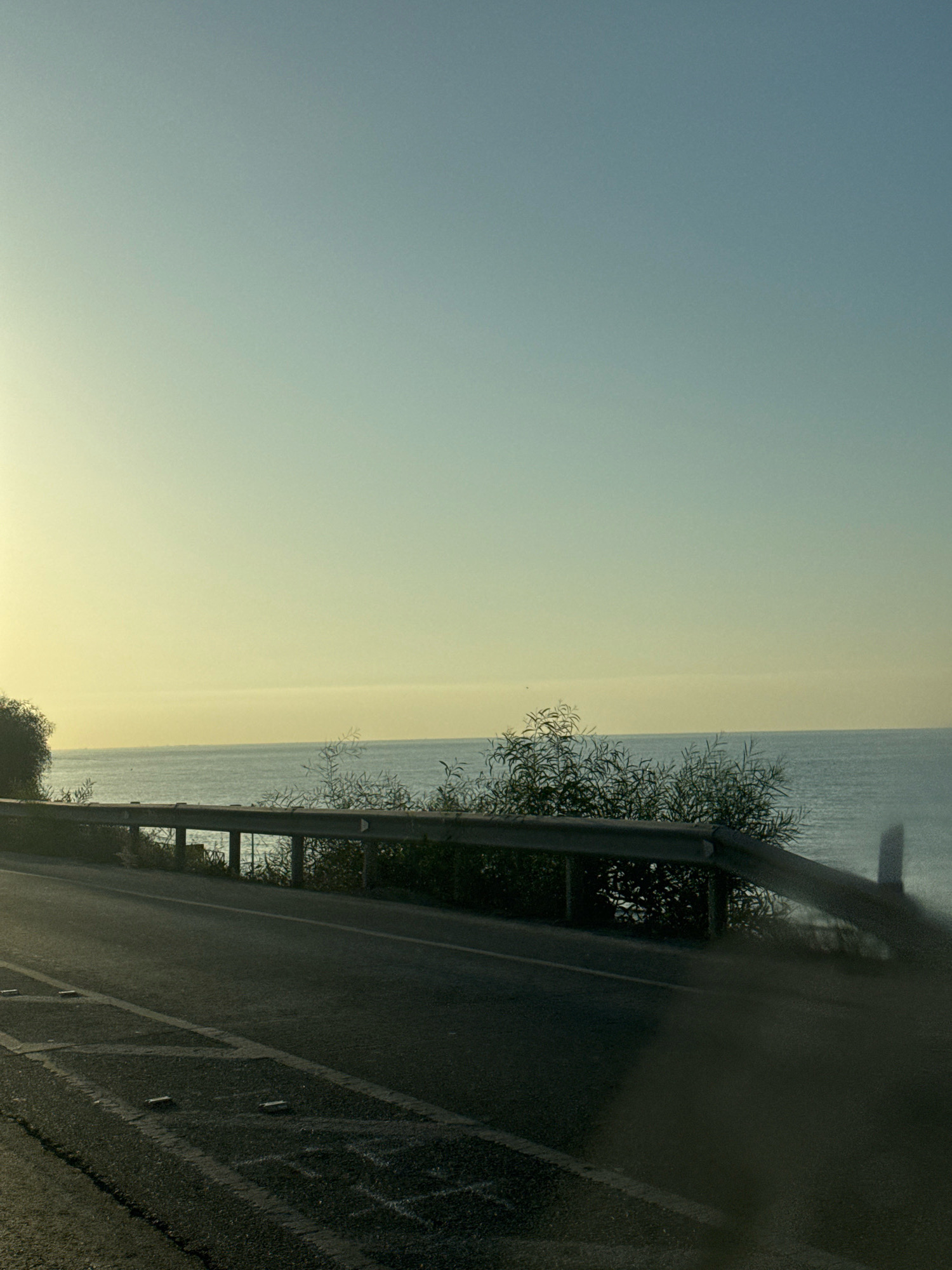 The image shows a scenic view of the sea from a coastal road in Agios Tychona Community, Cyprus. The road is paved and appears to be a two-lane highway with visible lane markings. A metal guardrail separates the road from the sea, which stretches out to the horizon under a clear, pale blue sky. Lush green vegetation, including trees and bushes, grows along the edge of the road and behind the guardrail, partially obscuring the view of the water. The sun is low in the sky, casting a warm, golden light, suggesting it might be sunrise or sunset. The overall atmosphere is serene and peaceful, capturing the beauty of the Cypriot coastline.