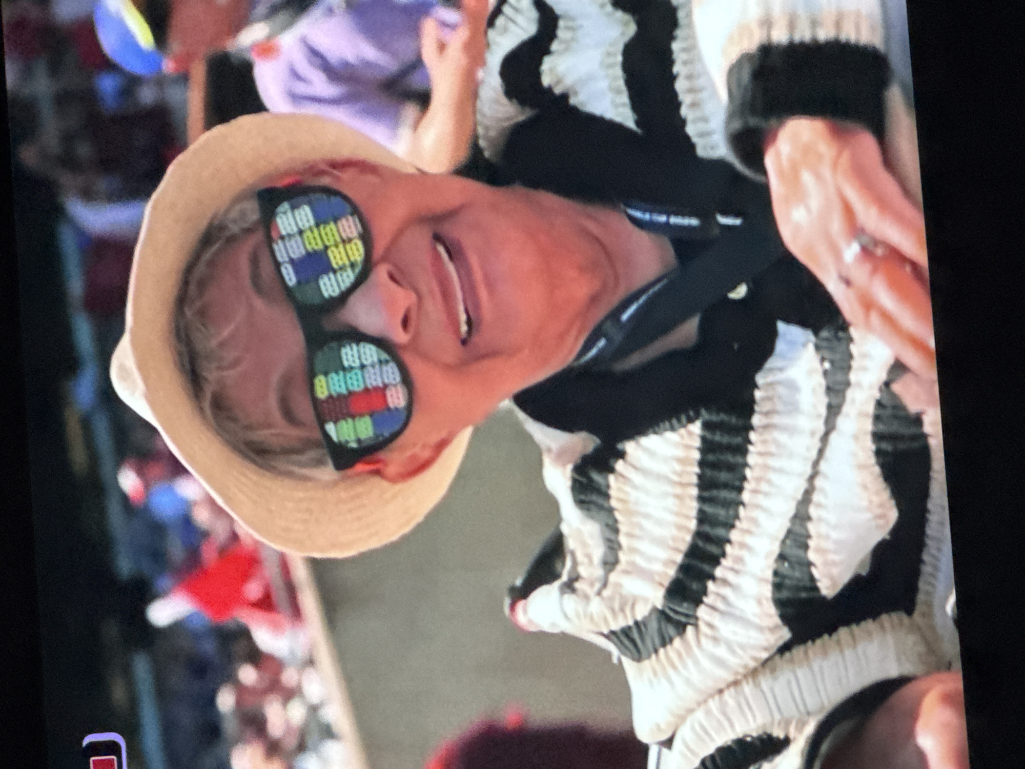 A woman with short blonde hair, wearing a straw hat and sunglasses, smiles at the camera. Her sunglasses reflect a colorful, pixelated pattern. She is wearing a black and white striped jacket with ruffled details. The background shows a crowd of people and a red flag. The image appears to be taken at an outdoor event, possibly a sporting event or festival.