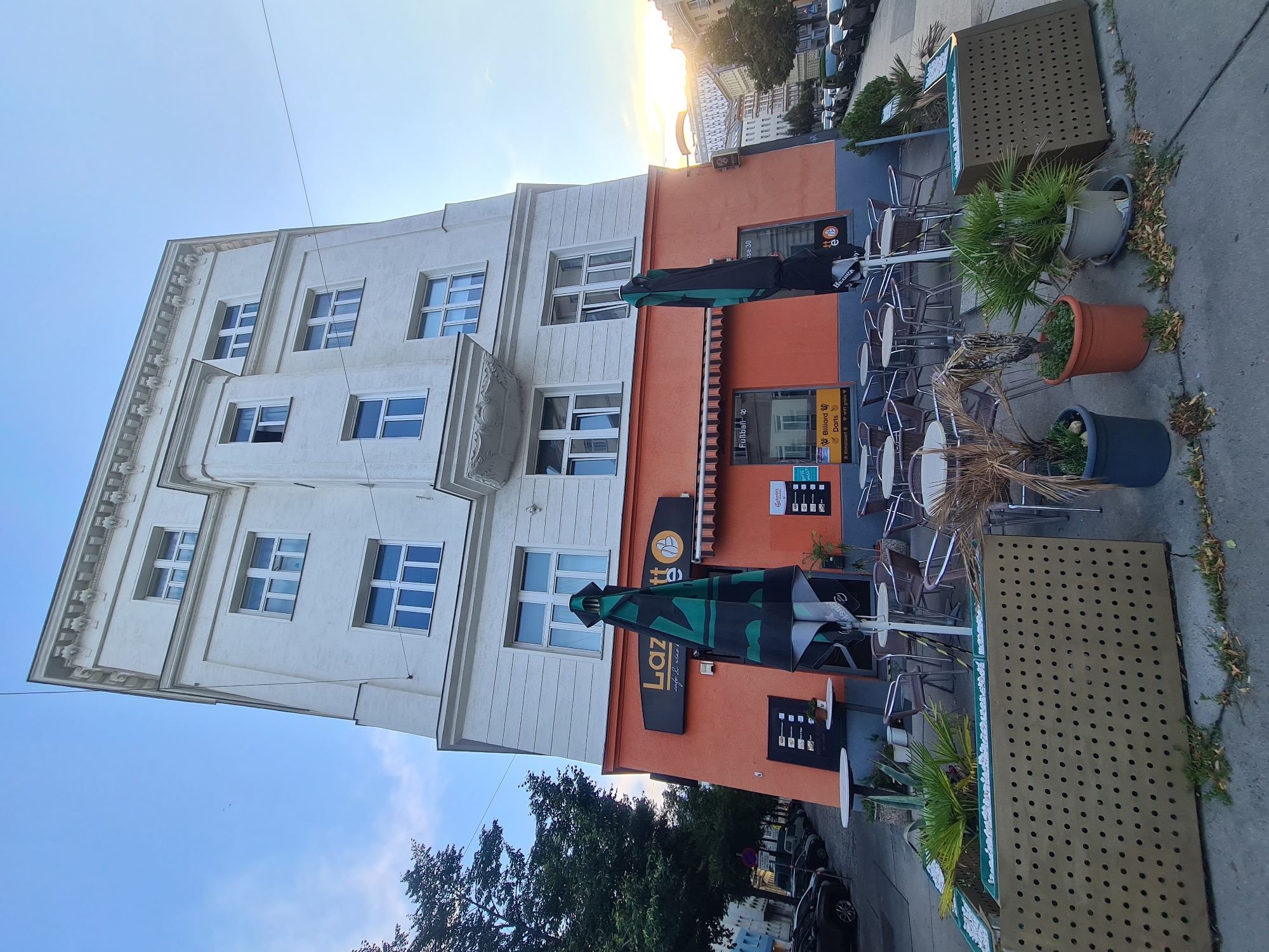 The image shows the facade of a multi-story building with an orange-colored ground floor that houses a cafe called "Lazarett Cafe". The cafe has outdoor seating with tables and chairs, and two large, closed green and black umbrellas are positioned in front of the entrance. A sign above the entrance displays "Lazarett Cafe" along with a logo featuring a coffee cup and a wheat stalk. Other signage on the orange facade advertises "Fußball", "Billiard Darts", "Klimatisiert", and "WiFi gratis". The building is situated on a street with parked cars visible along the sidewalk. The overall setting appears to be a European city street, with apartment buildings and a clear sky with some wispy clouds. The sun is setting, casting a warm glow on the scene and the buildings in the background. The ground floor cafe is likely in Vienna, Austria, given the presence of German language on the signs.