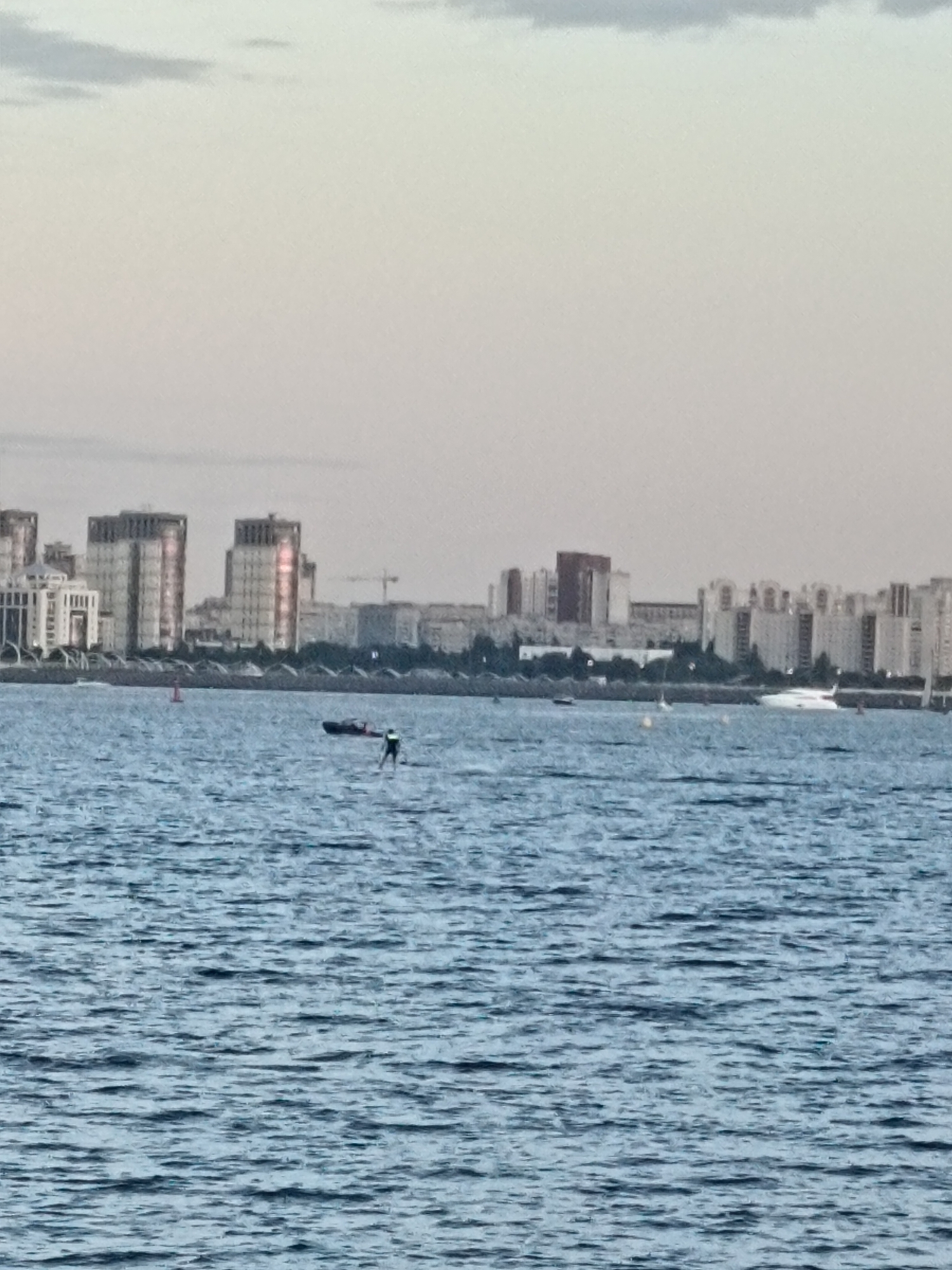 The image captures a wide shot of the Neva River in Saint Petersburg, Russia, with the city's skyline visible in the background. The water is choppy with small waves. In the middle ground, a person is stand-up paddleboarding. To the left of the paddleboarder, a dark-colored boat is visible. Further in the background, a white yacht is seen sailing on the right side. The city skyline is dominated by modern buildings, with some appearing to be residential complexes and others possibly office buildings. A crane indicates ongoing construction. The sky is overcast and a muted grey-white.