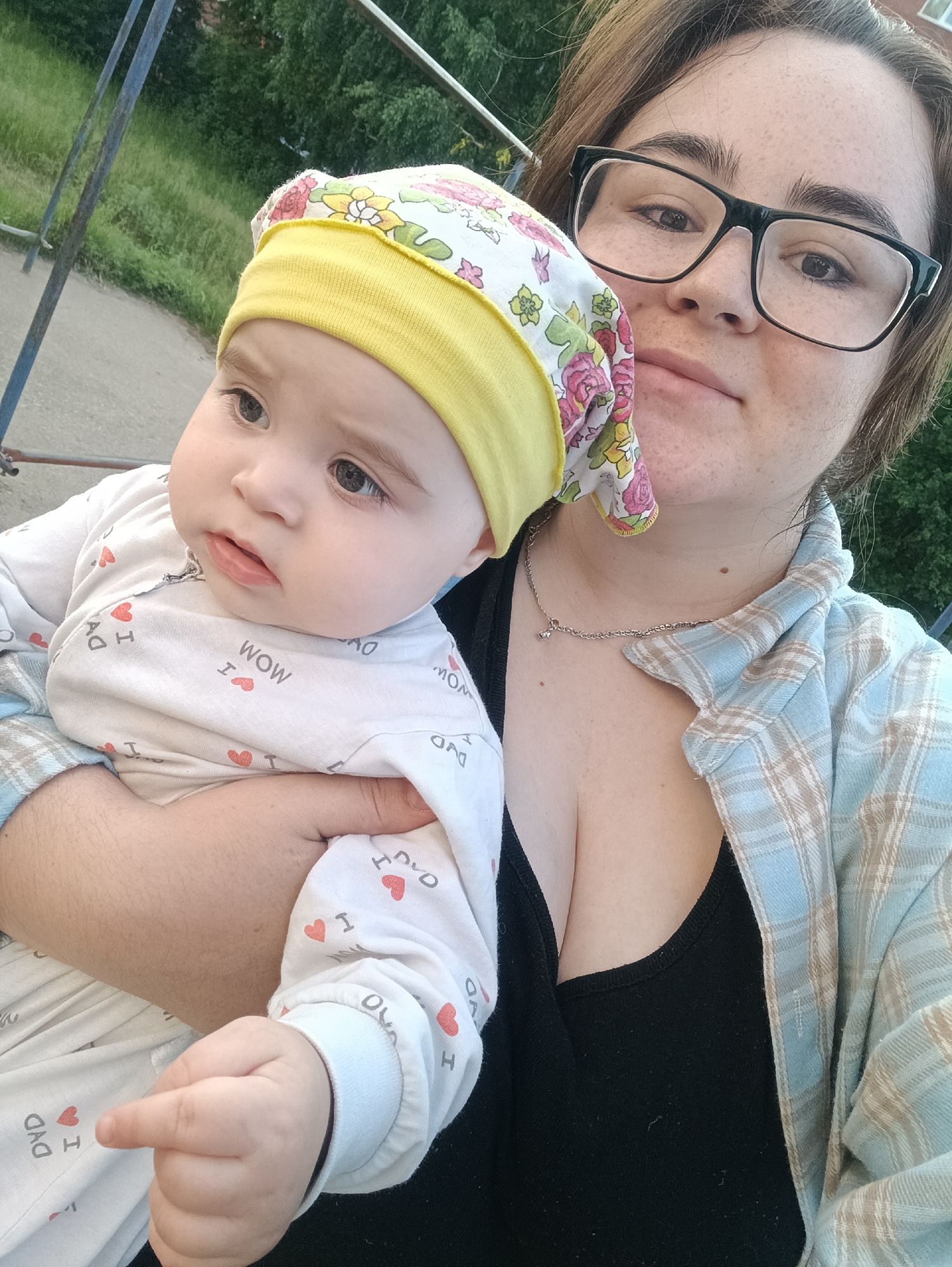 In an outdoor, possibly urban or suburban, environment with greenery and some play equipment in the background, a young woman with glasses is holding a baby. The woman is wearing a plaid shirt over a black tank top and a necklace. The baby is wearing a floral-patterned hat and a white onesie with red hearts and text. They are both looking towards the camera. The scene appears to be a casual, everyday moment.