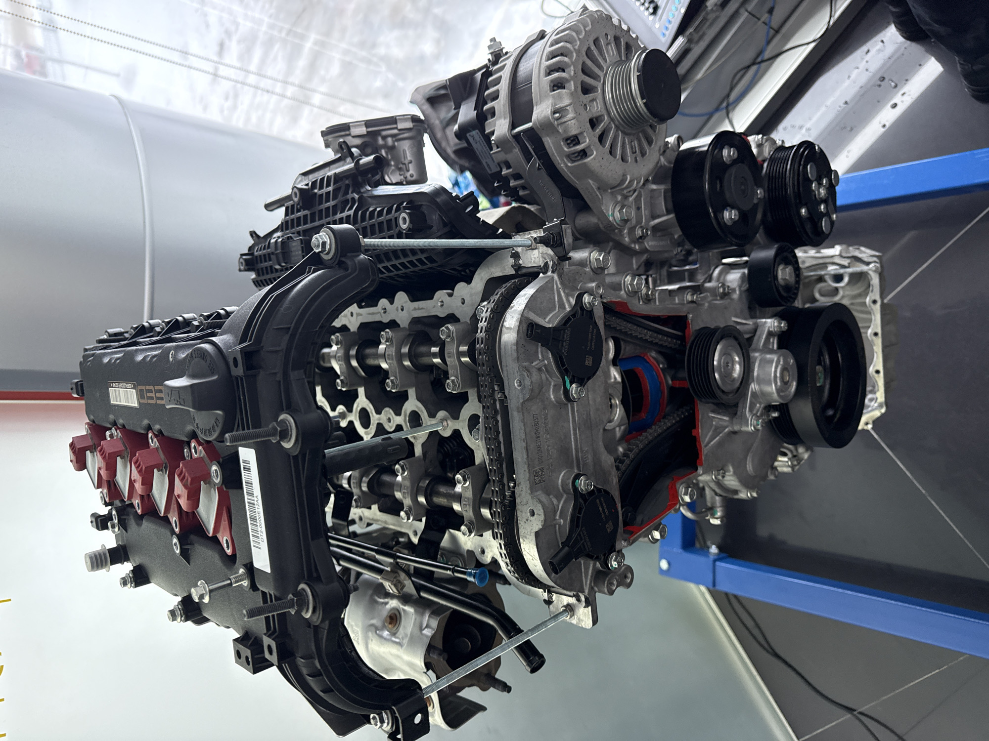 The image shows a partially disassembled engine mounted on a blue metal frame. The engine is in a laboratory or workshop setting, with gray metallic surfaces in the background. The left side of the engine is black and red, featuring components such as the valve cover and ignition coils. The right side reveals the internal components, including timing chains, belts, pulleys, and gears. An alternator is mounted on top. The image does not depict any people or indicate a specific location in Perm, Russia.