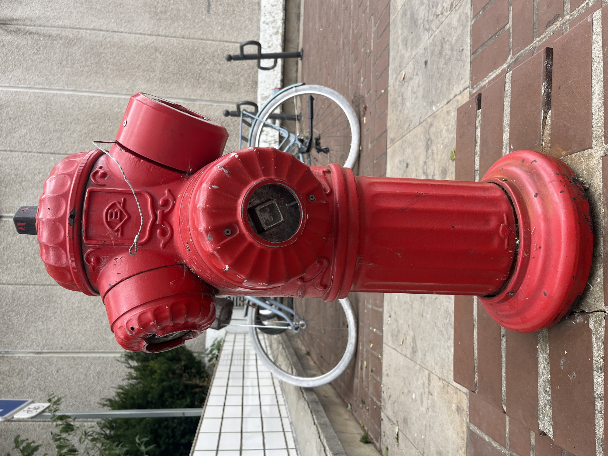 A red fire hydrant stands in the foreground on a sidewalk composed of brick and concrete paving stones. In the background, a light blue bicycle is parked, its wheels visible. Behind the bicycle, a textured concrete wall forms the backdrop. To the left of the bicycle, a white tiled wall runs horizontally, and further left, a green bush and a signpost with a blue and white symbol are visible. The scene appears to be an outdoor urban environment.
