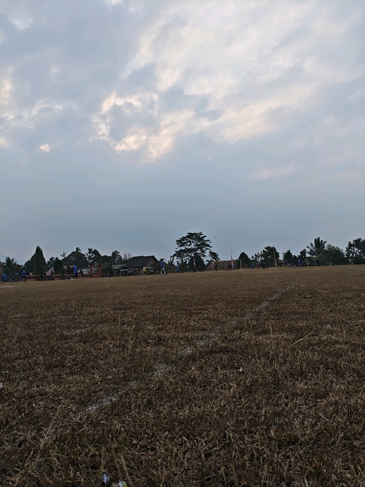 The image depicts a dry, brown soccer field with several individuals in blue uniforms playing a match. The field is largely devoid of green grass, indicating a dry season. In the background, a line of trees separates the field from a cluster of buildings, suggesting a semi-rural or residential area. The sky is overcast with soft clouds, and the overall lighting suggests it is late afternoon or early evening. There are goalposts visible on the field. The scene appears to be set in Karang Sari, Indonesia, based on the provided context.