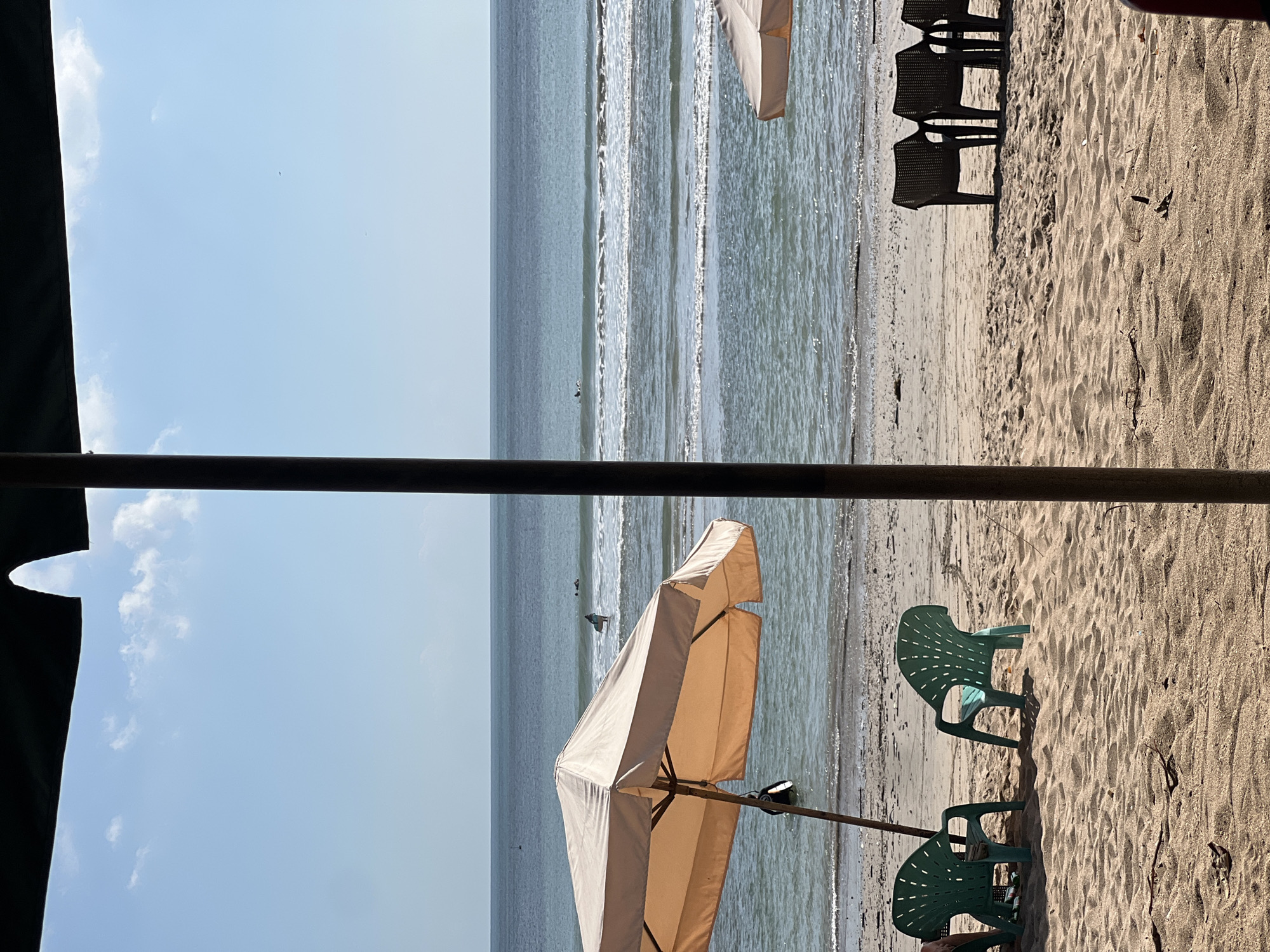 In Kuta, Indonesia, a tranquil beach scene unfolds under a clear blue sky with scattered clouds. The foreground is dominated by golden sand, lightly scattered with dried leaves and twigs. Two empty, bright green plastic beach chairs stand side-by-side on the sand, casting distinct shadows. To the right, a set of dark brown woven plastic lounge chairs are arranged in a row, also unoccupied.
A large, light tan beach umbrella, supported by a wooden pole, extends over the left side of the frame, providing shade over the green chairs. Part of a dark structure, possibly a pillar or a beam, is visible on the far right and left edges of the image, framing the view.
Beyond the sand, the ocean presents a gradient of blues and greens, with gentle waves rolling towards the shore. Several surfers are visible in the water, some riding waves and others waiting. A person is standing in the shallow water near the umbrella, holding a surfboard. In the distance, the horizon line separates the ocean from the sky, with a thin strip of land visible. Another beach umbrella, partially obscured, is situated on the far right, closer to the water's edge.