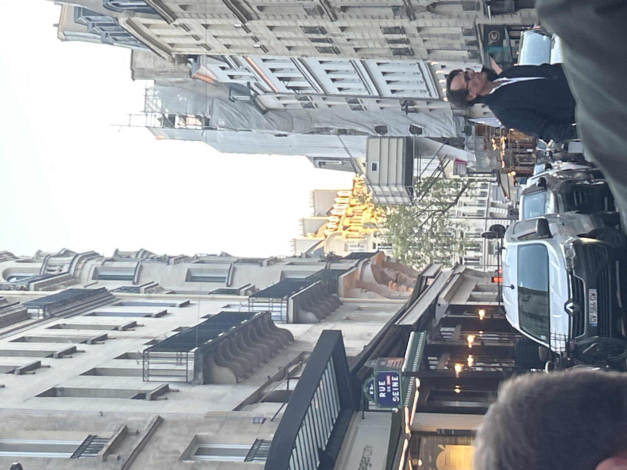 The image depicts a street scene in Paris, France. The street is lined with buildings, mostly constructed of beige stone, with ornate balconies and detailing. A sign that reads "Rue de Seine" is visible on the building at the street corner. Several vehicles are parked on the street, including a white van and dark colored cars. A man wearing a dark jacket and glasses is standing on the sidewalk. Construction is occurring on a building in the background. The photograph is taken at street level.