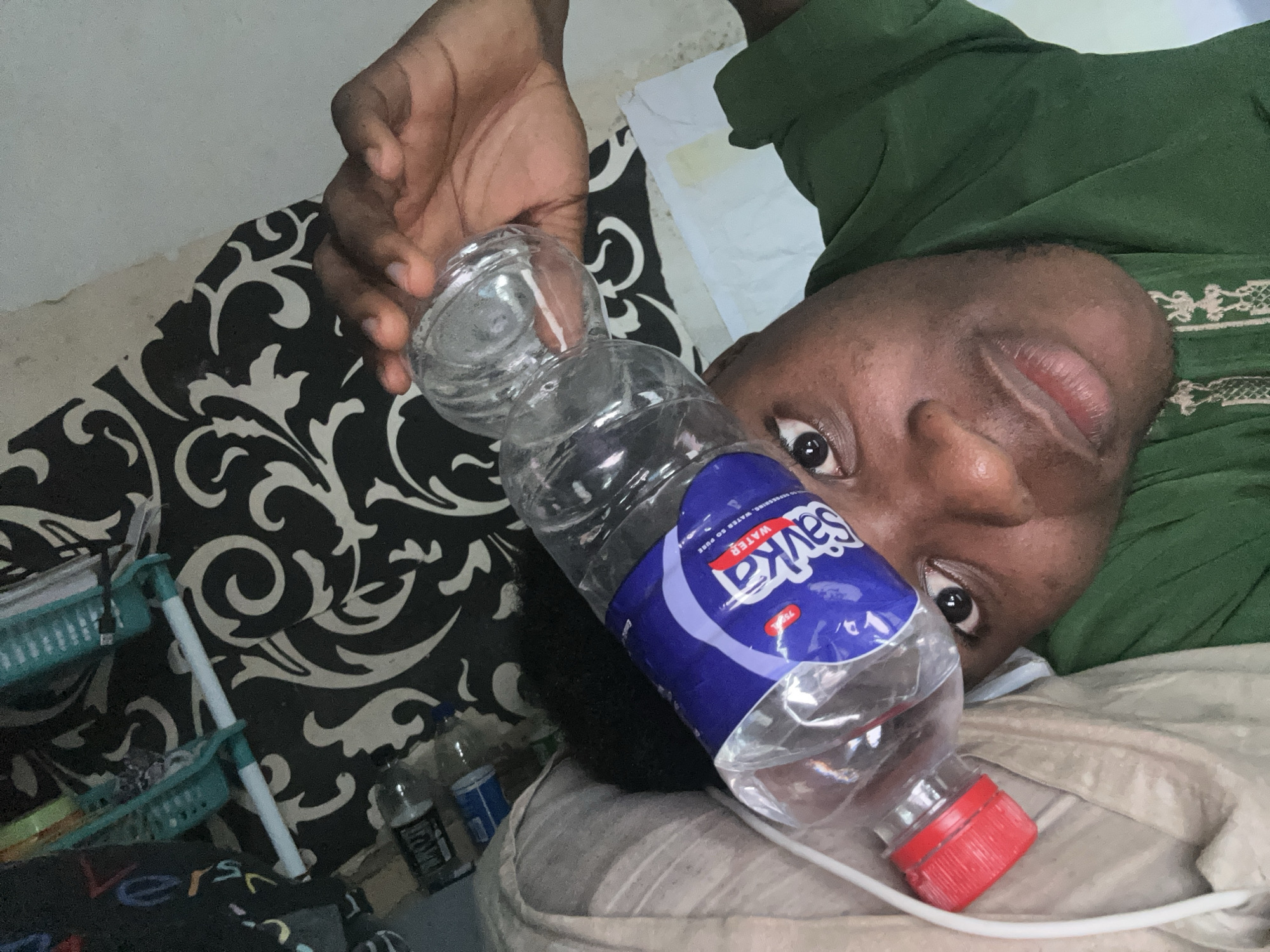 The media file is a selfie taken by a person lying down, likely in a bedroom given the presence of bedding and a patterned fabric. The person has dark skin and is looking towards the camera with their eyes open. They are wearing a green garment, possibly a thobe or abaya, which is draped over them.
In the foreground, a partially full plastic water bottle with a red cap is held by a hand. The bottle has a blue and white label with the brand name "Savka WATER" clearly visible. The text on the label also includes "WATER SO PURE" and "750 ml".
In the background, there is a black and white patterned fabric, possibly a bedsheet or a decorative cloth. Next to it, there are two teal-colored plastic baskets, one on top of the other, containing various items like pens or small tools. There are also other water bottles visible in the background.
The lighting suggests it is daytime, though it is difficult to determine the exact time of day or weather conditions. The overall mood appears relaxed, perhaps the person is resting. The location context provided is Ihiagwa, Nigeria, which aligns with the appearance of the person and the general domestic setting.