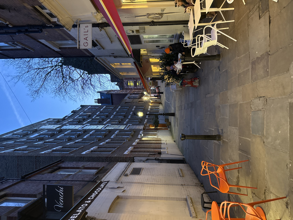 A narrow European street scene at dusk. Buildings line both sides of a cobblestone path, lit by street lamps. A "Gail's" sign is visible on the left. Tables and chairs, some white and some orange, are placed along the street for outdoor seating. A "Venchi Gelato & Chocolate" sign is seen on the right. A person wearing an orange hat is seated at a table. Bare tree branches reach into the evening sky above the buildings.