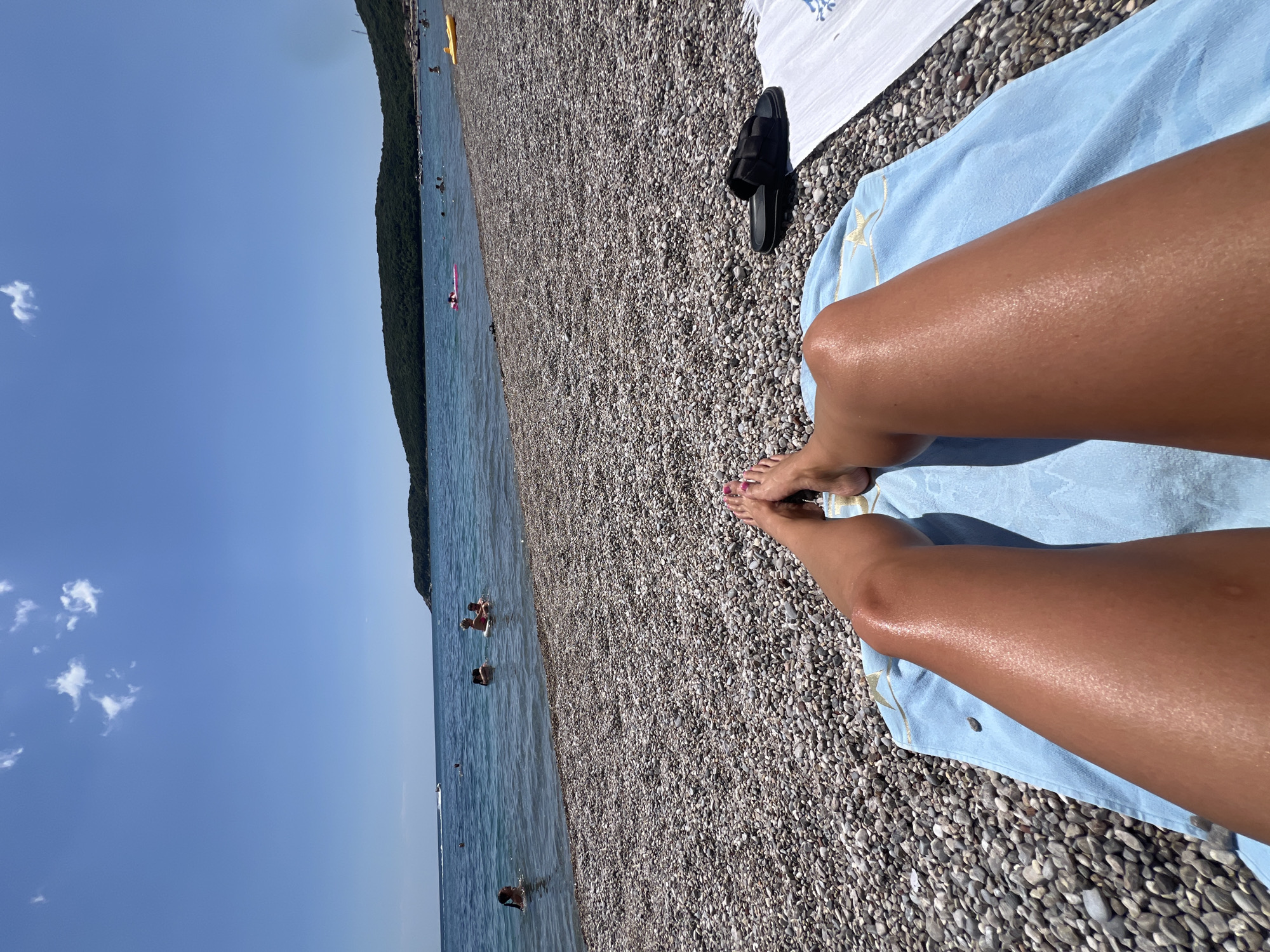 The image depicts a sunny scene at a beach in Prijevor, Montenegro. In the foreground, a person's legs are resting on a light blue towel placed on a pebble beach. A pair of black sandals are visible near the towel. In the background, there is a view of the Adriatic Sea with a few people swimming. A vegetated coastline is visible along the horizon. The sky is clear blue with a few scattered clouds.