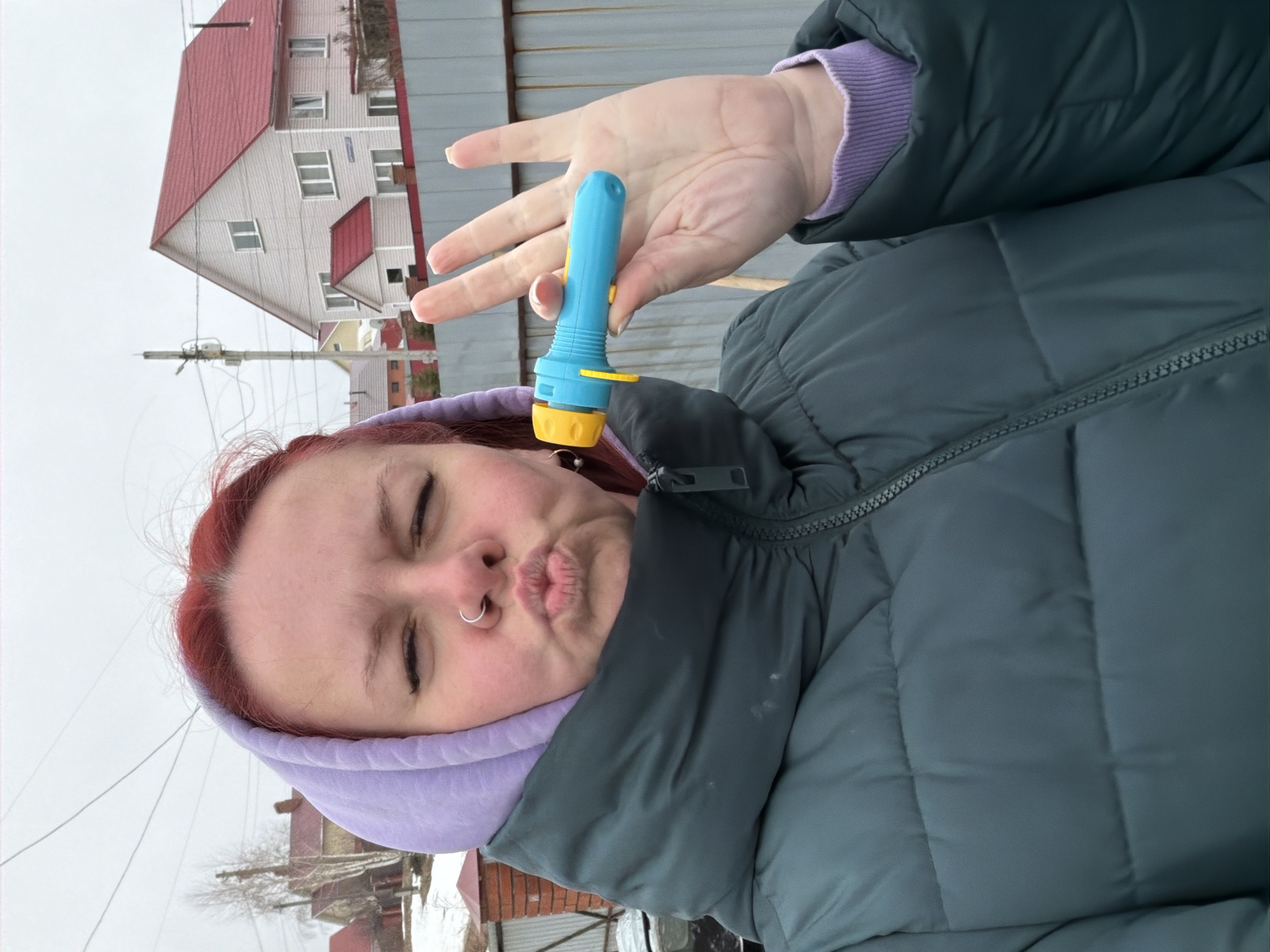 A woman is holding a blue toy flashlight. She has red hair and a nose ring. She is wearing a purple hoodie and a gray puffer jacket. The background shows a residential area, including a house with a red roof. The location is identified as Chaikovsky, Russia.
