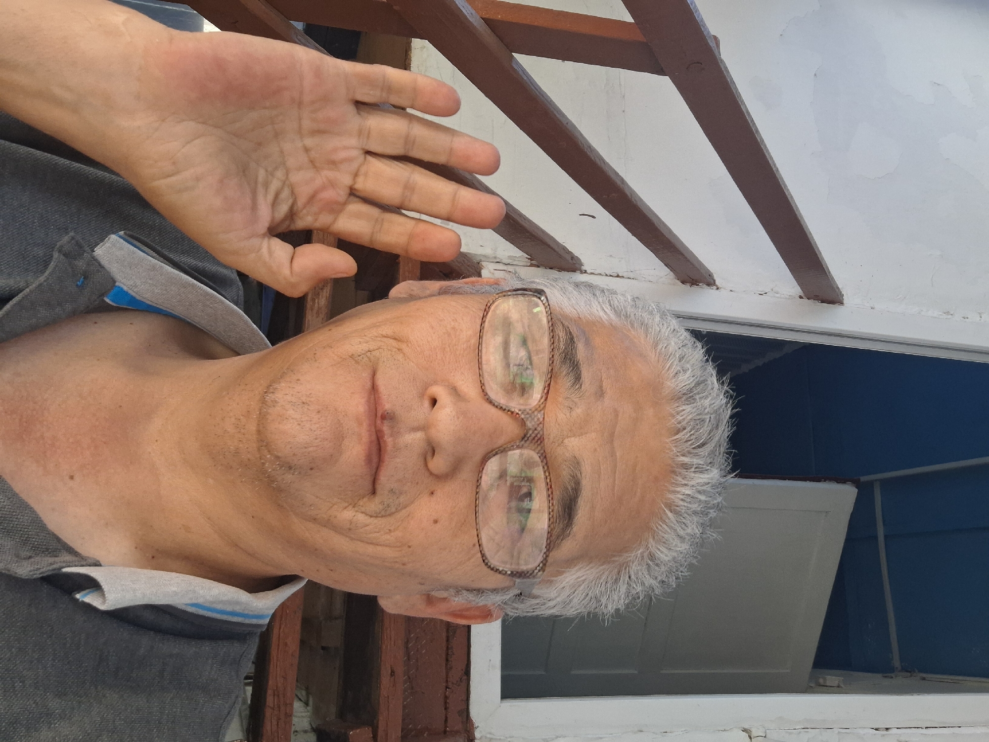 A selfie captures an older East Asian man with grey hair and glasses, wearing a grey polo shirt. He is standing outdoors in front of a white building with wooden railings. His right hand is raised and open, with his palm facing the camera, in a waving gesture. The background shows wooden stairs leading up to a doorway. The lighting suggests it is daytime. The location is Almaty, Kazakhstan.