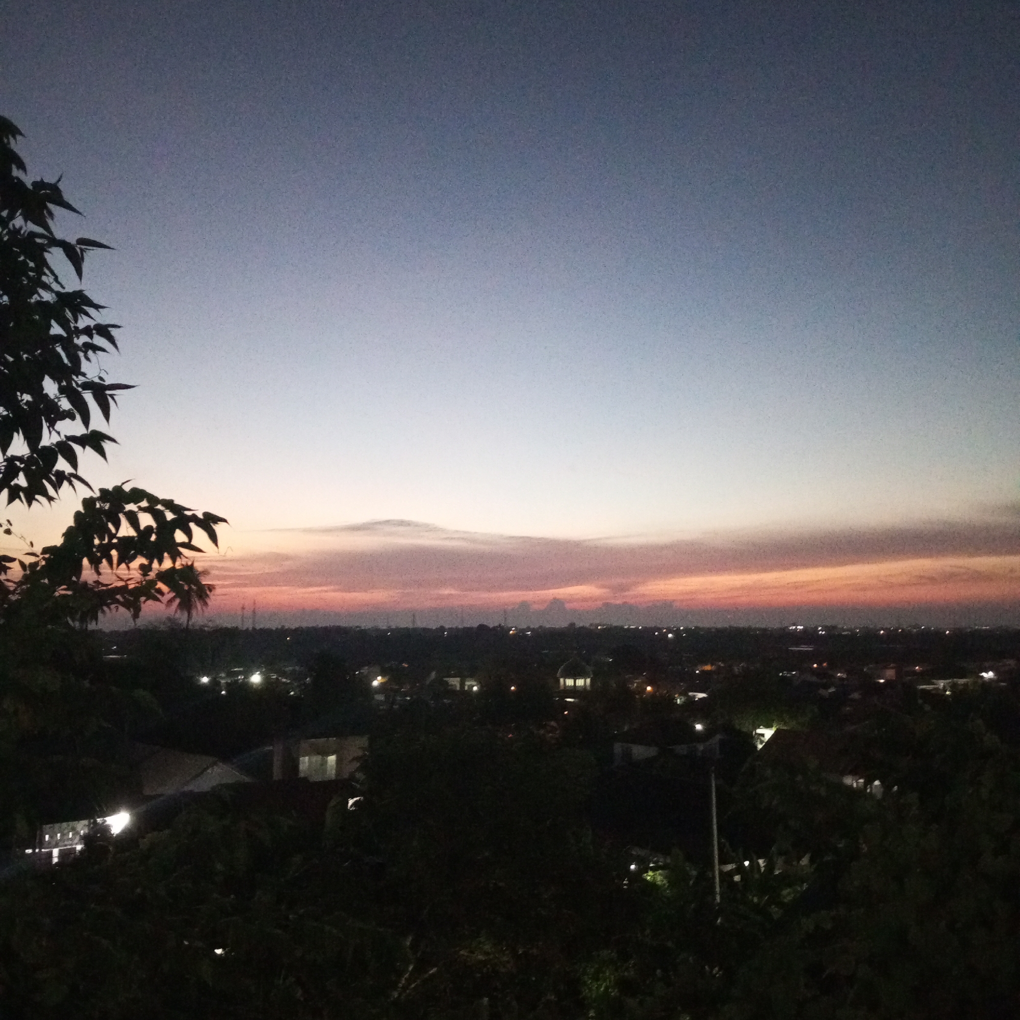 An outdoor, high-angle shot shows the evening sky over Lhokseumawe, Indonesia. The sky has a gradient from deep blue at the top to pale yellow near the horizon, with thin bands of orange and pink in between. The urban landscape below is mostly obscured by trees and foliage in the foreground, but scattered lights from buildings and streetlights are visible, suggesting a residential area. A silhouette of tree branches occupies the left side of the frame.