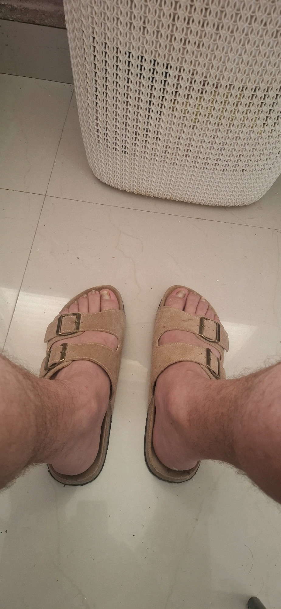 A first-person perspective shot shows a person's feet wearing tan Birkenstock sandals, standing on a tiled floor. Behind the feet is a white woven laundry basket. The scene appears to be indoors, likely in a residential setting. There is no indication of the location being in Yerocham, Israel.