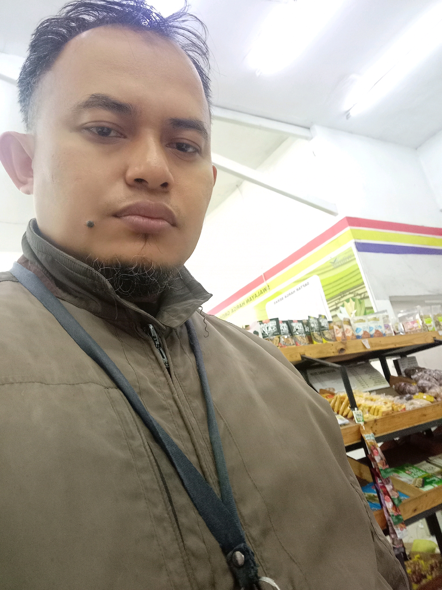 The image is a selfie taken indoors, likely in a convenience store or market. The subject is a man with short, dark hair styled with a fade. He has a mole on his chin and a light goatee. He is wearing a brown zippered jacket with a dark blue lanyard around his neck. In the background, shelves are visible displaying various food items and packaged goods. A sign with colorful stripes and text, possibly in Indonesian, is also present. The lighting suggests an indoor environment with overhead fluorescent lights. The setting appears to be within a commercial establishment in Mulyoagung, Indonesia, as indicated by the signage and typical store layout.