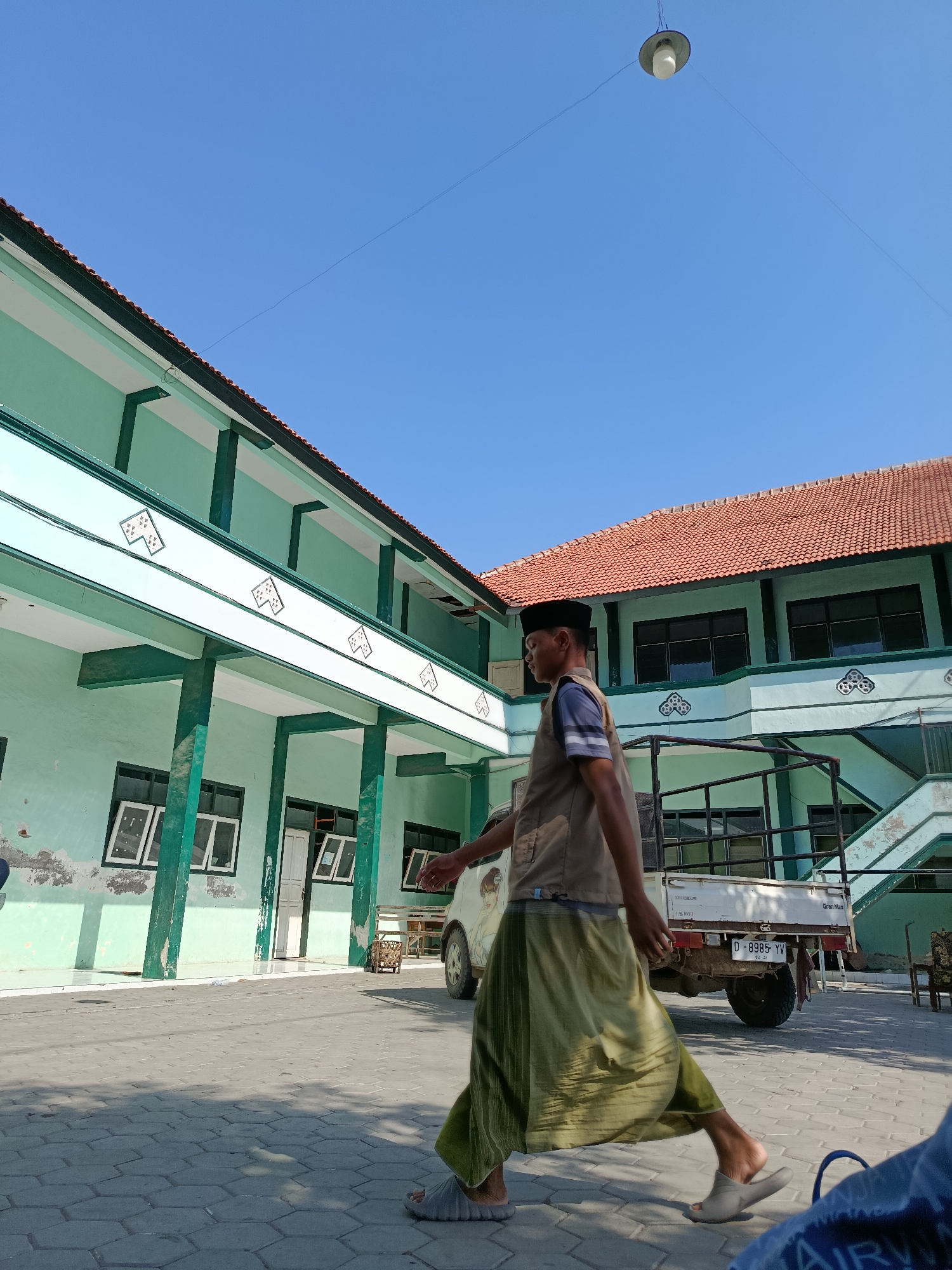 A man wearing a peci hat, a striped shirt, a vest, and a sarong walks across a paved courtyard in front of a two-story building with a red tile roof. The building is painted pale green with white trim. The man is walking from left to right in the frame, with his body angled slightly away from the camera. A white pickup truck with a license plate that reads "D 8985 YV" is parked in the background to the right of the man. The sky is clear and blue. The scene appears to be in Ramban Wetan, Indonesia.
