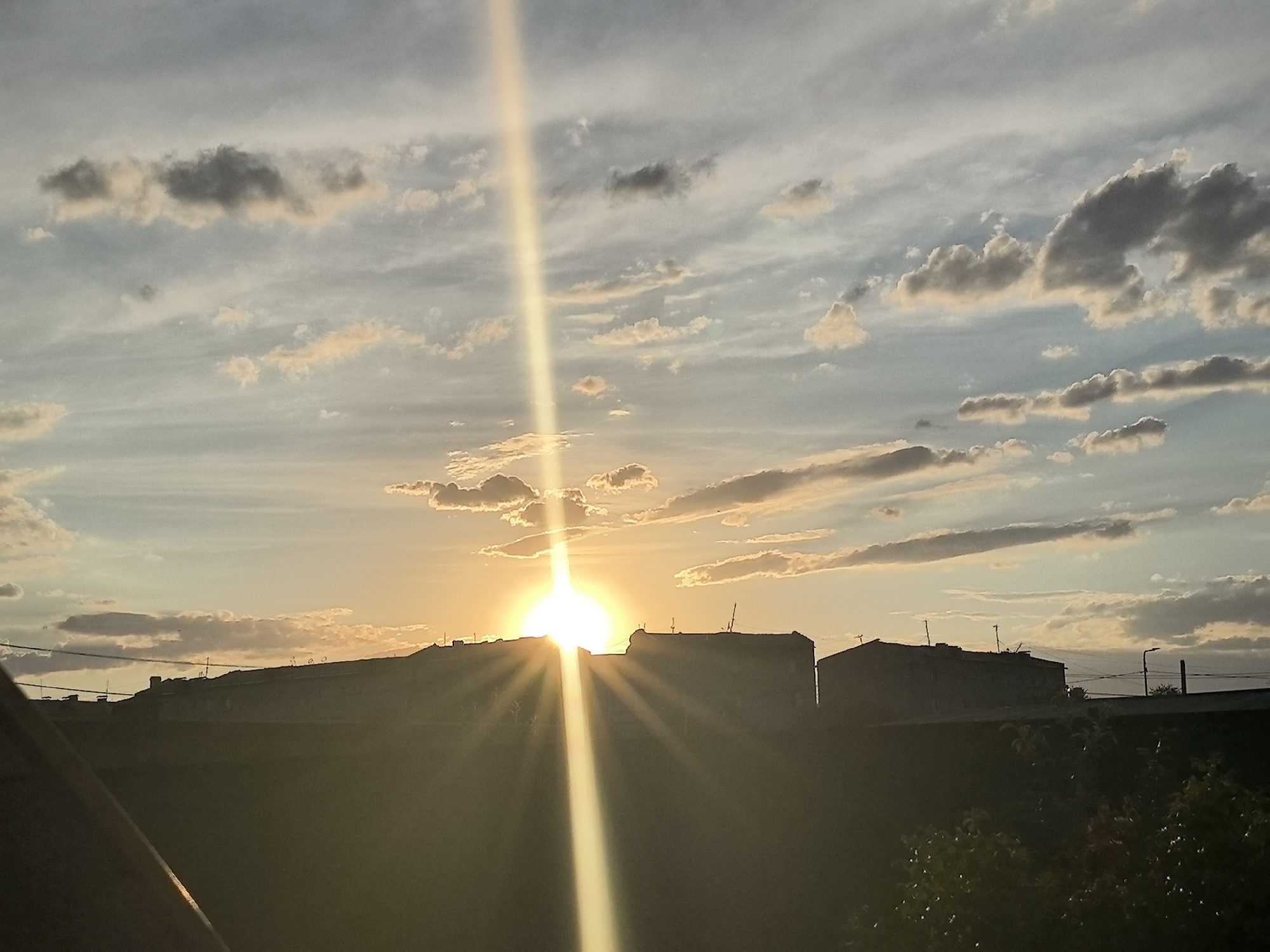 A low-angle, wide shot captures a sunset over the city of Taldykorgan, Kazakhstan. The sun, a bright orb, is partially obscured by a silhouetted building on the horizon, casting brilliant rays across the sky. Wispy clouds, tinged with orange and yellow from the setting sun, drift across a pale blue sky. The foreground is dominated by a dark, silhouetted foreground, possibly a rooftop or balcony, with foliage visible in the lower right corner. In the mid-ground, the outlines of buildings and utility poles are visible against the illuminated sky. The overall scene evokes a peaceful, end-of-day atmosphere.