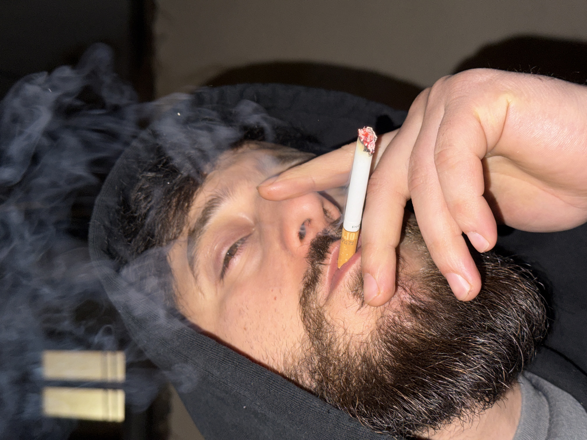 A close-up, angled portrait features an adult male subject in Strasbourg, France, smoking a cigarette. The individual possesses dark hair and a full brown beard, and is wearing a black hooded sweatshirt with the hood drawn over his head. He is holding a lit cigarette, which has an orange filter, between the thumb and index finger of his right hand, positioned near his mouth. Visible smoke emanates from the cigarette and ascends around his head. His eyes are partially closed. The background is dark and indistinct, with two blurry, vertical, light-colored rectangular objects discernible in the upper left quadrant. The scene appears to be dimly lit.