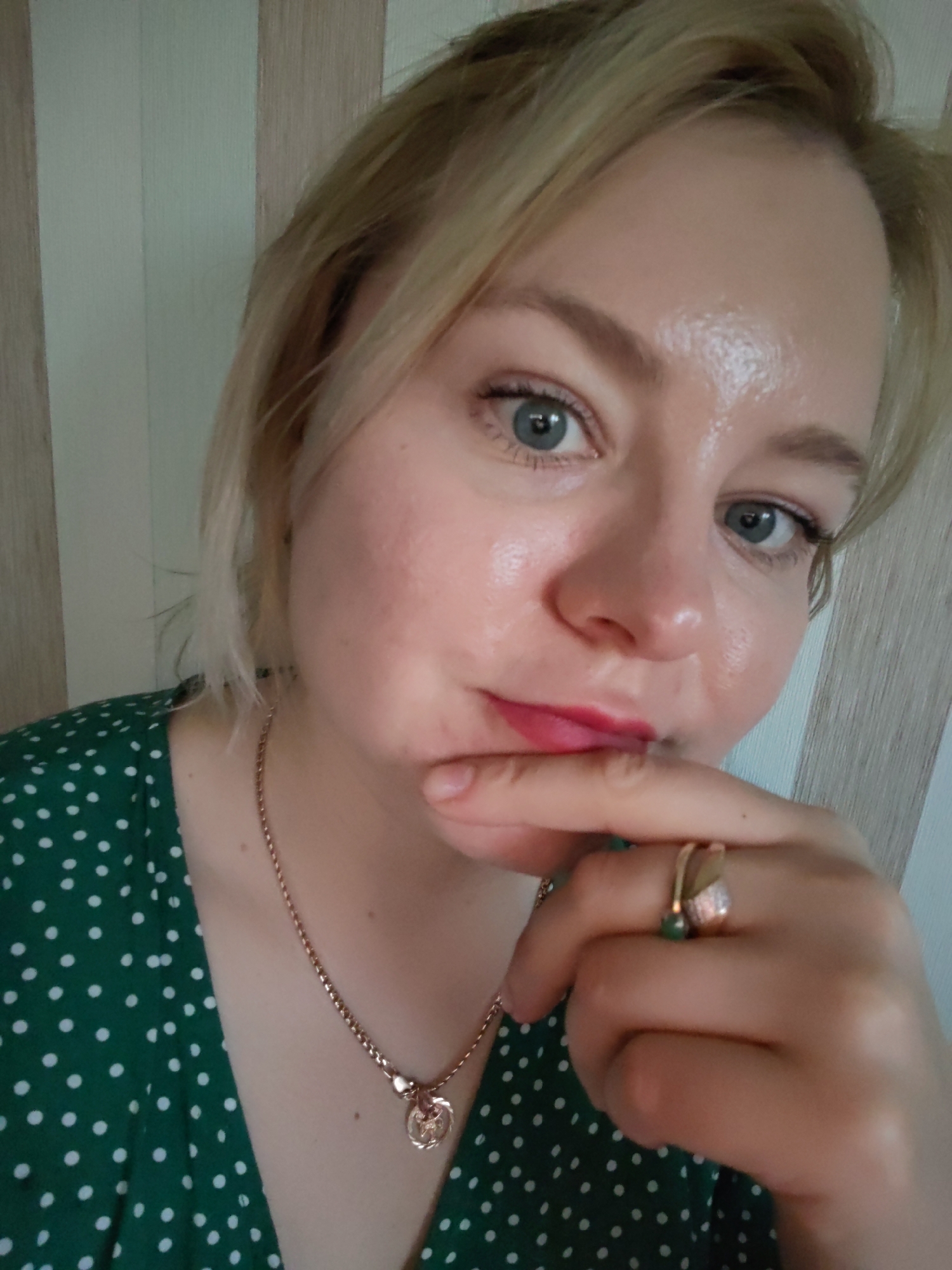 A close-up portrait features a fair-skinned woman with short, light blonde hair. She looks directly at the camera with wide, light blue-grey eyes, her right hand raised to her chin with a finger resting on her lower lip, conveying a thoughtful or slightly pensive expression. She is wearing a dark green top with white polka dots. A delicate gold-colored chain necklace with a small, round charm hangs around her neck. On her right hand, two rings are visible: one with a prominent green stone and another with small, clear stones on a gold-colored band. She has red lipstick on. The background features a light-colored wall with vertical stripes, suggesting an indoor setting. The lighting is soft and even, illuminating her face, which has a natural sheen. There is no visible text in the image.