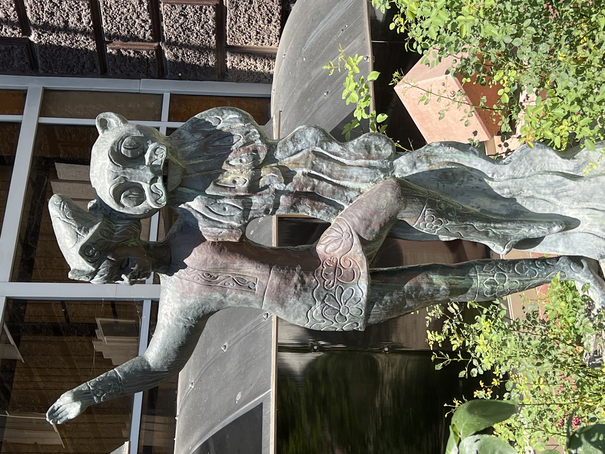 This media file shows a bronze sculpture of a fantastical creature with a feline head and human-like body, in a garden setting. The sculpture appears to be made of patinated bronze, with areas of green and brown. The creature's head is large and stylized, with prominent ears, eyes, and a mouth. It has a flowing mane or headdress. The body is slender and appears to be in motion, with one arm extended forward and the other bent at the elbow. The legs are also positioned as if in mid-stride, with decorative patterns on the leggings.
Behind the sculpture, there is a building with a textured brown facade and large glass windows. Green foliage from a plant, possibly a rose bush, is growing in the foreground and around the sculpture, obscuring parts of the background. The scene is brightly lit, suggesting it is daytime, and the sunlight casts shadows on the sculpture and the building. The setting appears to be an outdoor public space, possibly a park or a garden. There are no visible text elements in the image. The overall impression is one of artistic expression and nature integration.