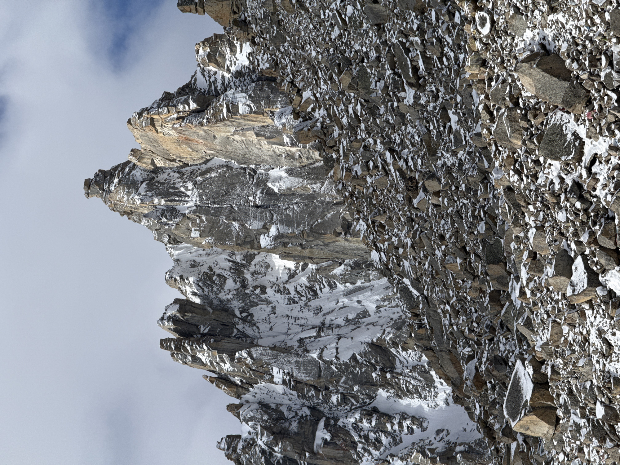 The image displays a rugged and imposing mountain or cliff face, composed primarily of sharp, fragmented rocks in shades of grey and brownish-grey. Large areas of the rock are covered with white snow and ice, clinging to ledges, cracks, and rough surfaces, indicating a cold, high-altitude environment. The steep terrain features a dramatic interplay of light and shadow, highlighting the intricate textures and numerous angular rock formations.
The upper left portion of the image reveals a light-grey sky with some scattered clouds and hints of blue. The bright daylight illuminates the scene, enhancing the stark contrast between the dark rock and pristine snow. There are no people, animals, or man-made objects visible, suggesting an untouched natural landscape. No specific activities or events are taking place; the scene is static and serene. The overall impression is one of grandeur and rugged wilderness.
No visible text is present in the media. This mountainous scene is situated near Parga town, China.