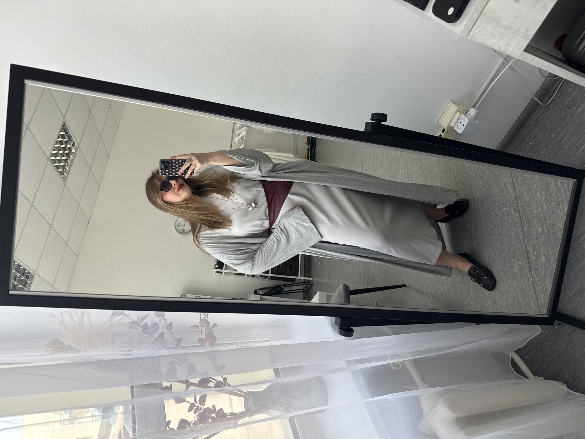 A woman is reflected in a large mirror with a black frame. She is taking a selfie with a smartphone that has a black and white polka dot case. She is wearing sunglasses and a long, gray dress with a burgundy detail, and a gray cardigan. Black shoes are visible on her feet.
The reflection in the mirror shows an interior space with light-colored walls and a patterned ceiling. Various pieces of furniture, including a rack with clothing, a chair, and shelves, are visible. A clock hangs on the wall in the background.
The foreground of the image includes a sheer, white curtain that partially obscures a window. The location is Izhevsk, Russia.