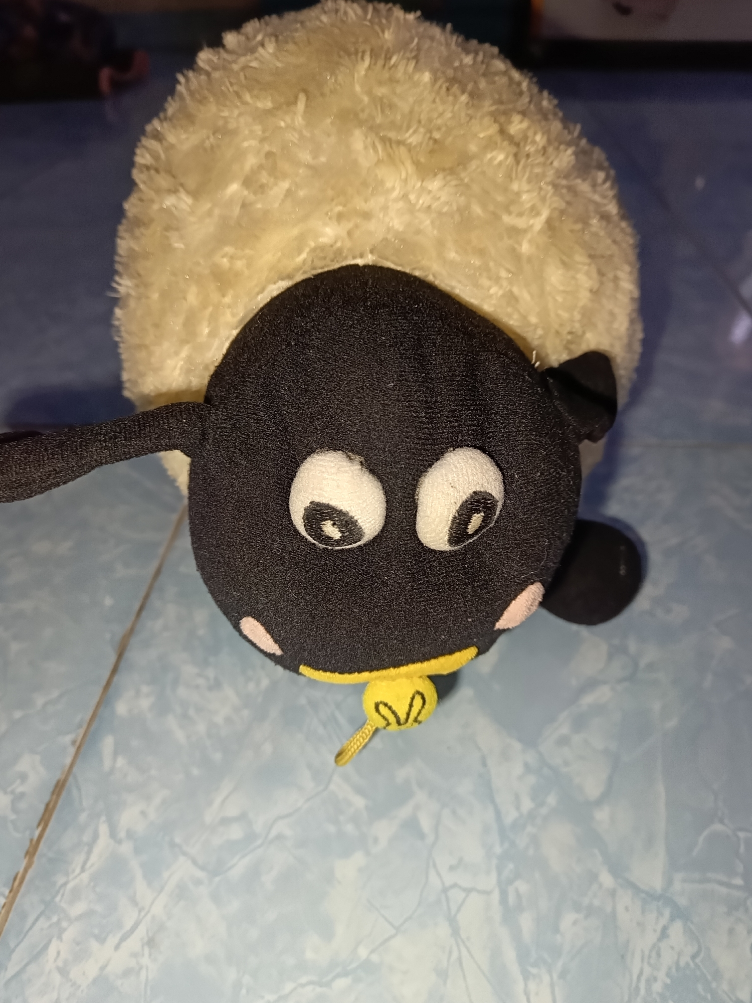 A stuffed sheep toy is positioned on a light blue tiled floor. The toy has a fluffy, cream-colored body and a black head with large, white eyes that have black pupils. It has two small, pink blush marks on its cheeks and a yellow collar with a small, yellow charm. The background is blurry and appears to be an indoor setting with some dark objects visible. The scene is well-lit, and the focus is on the toy. The location is not specified in the image.