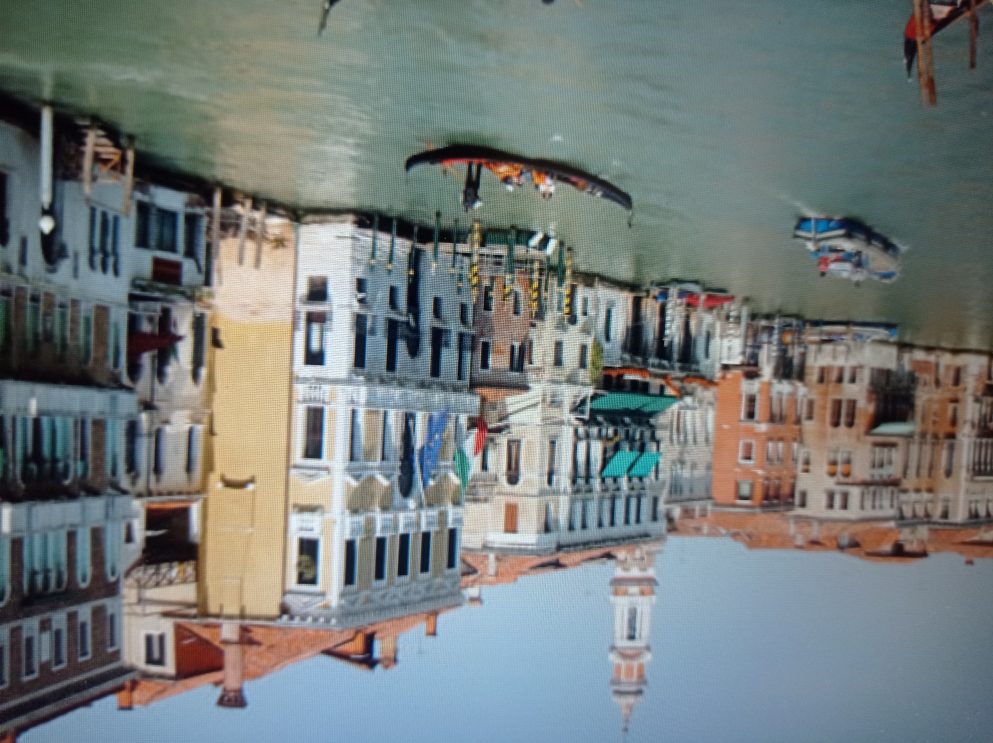 This is a slightly pixelated, high-angle, long-shot image of a canal in Venice, Italy. The canal is bordered by tall, colorful buildings with terracotta roofs. Several boats are visible on the water. In the foreground, a gondola carries several passengers and a gondolier poles it through the water. Further in the distance, a blue and white barge navigates the canal, also with people on board. The sky is a clear, pale blue. The buildings appear to be a mix of residential and commercial structures, with many windows and balconies. The architecture is classic Venetian, with ornate details. The overall scene depicts a typical day in Venice, with its iconic waterways and historic buildings. There is no indication that this image is related to Mukachevo, Ukraine.