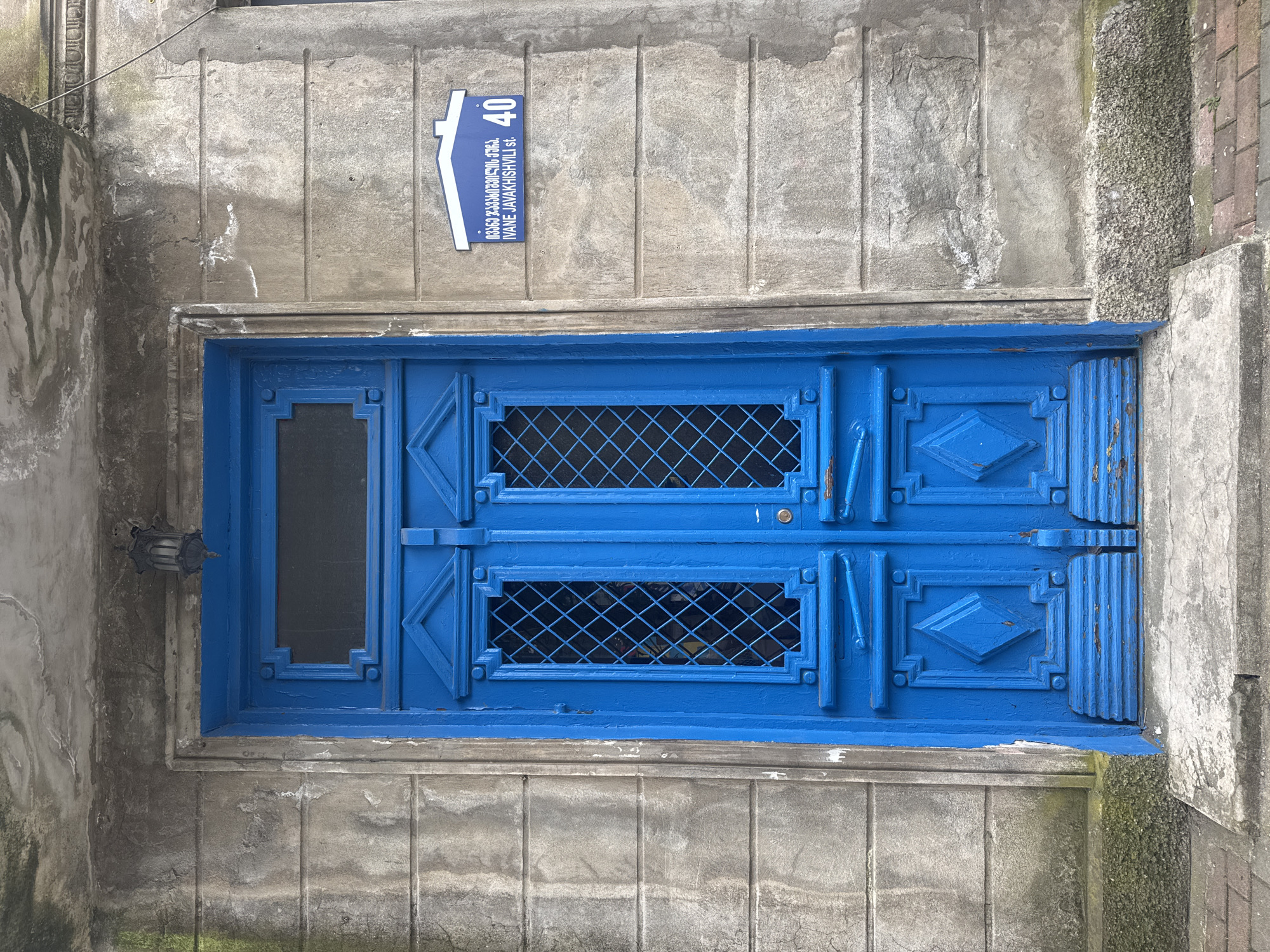 A blue door with a small window and metal grates is set in a gray, weathered wall. Above the door, there is a street sign with the number "40" and text in Georgian and English that reads "IVANE JAVAKHISHVILI st." The door appears to be located in a city. The city and country is unknown.