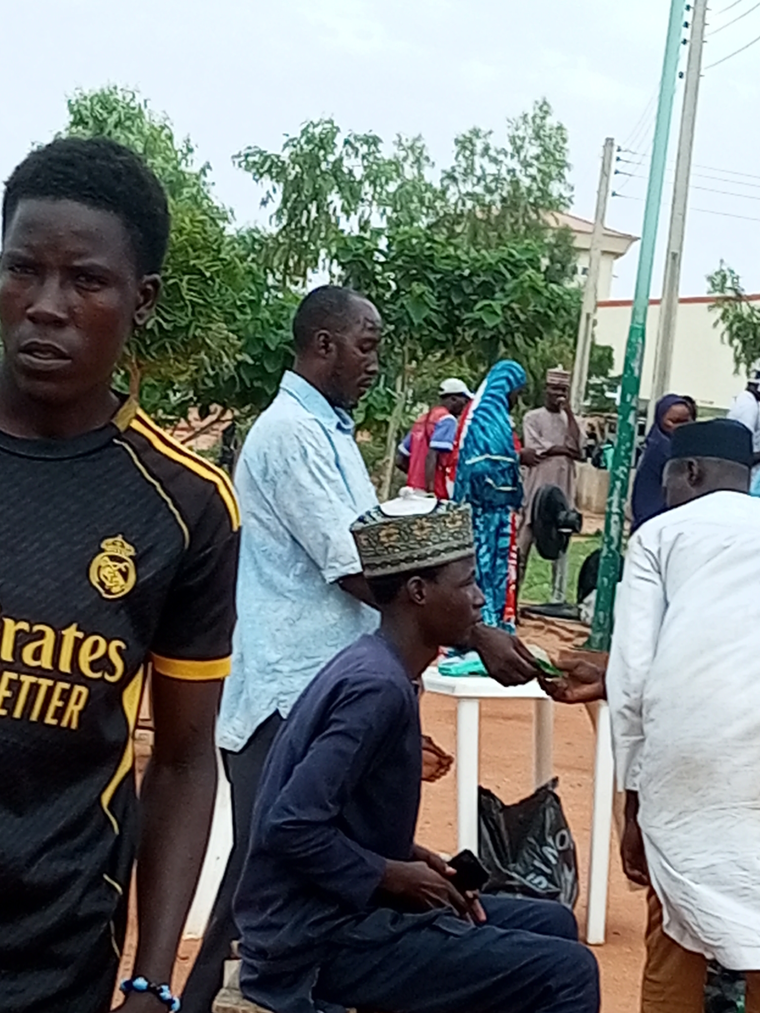 The image depicts an outdoor gathering in Gombe, Nigeria, characterized by a moderate overcast sky and lush green trees in the background. In the foreground on the left, a young man wearing a black and yellow football jersey with the "Emirates" and "Better" logos is prominently featured, looking towards the left. Adjacent to him, a man in a light blue shirt is seen from the side, facing away from the camera.
In the center, a man wearing a dark blue traditional outfit and a patterned cap is seated and appears to be interacting with another person whose hands are extended towards him. The interaction involves the exchange of what appear to be thin, green objects, possibly currency or documents. To the right of this group, a woman in a blue and white patterned hijab is standing, and further back, several other individuals are visible, including a man in a light brown outfit and a woman in a dark hijab.
A white plastic table stands in the middle ground, with a black plastic bag placed beneath it. A black electric fan is positioned behind the table. The ground is a sandy or dirt surface. A utilitarian pole with electrical wiring runs vertically on the right side of the image, and a building with a light-colored facade and an orange roof is visible in the background. The overall scene suggests a community gathering or event taking place in an open area.