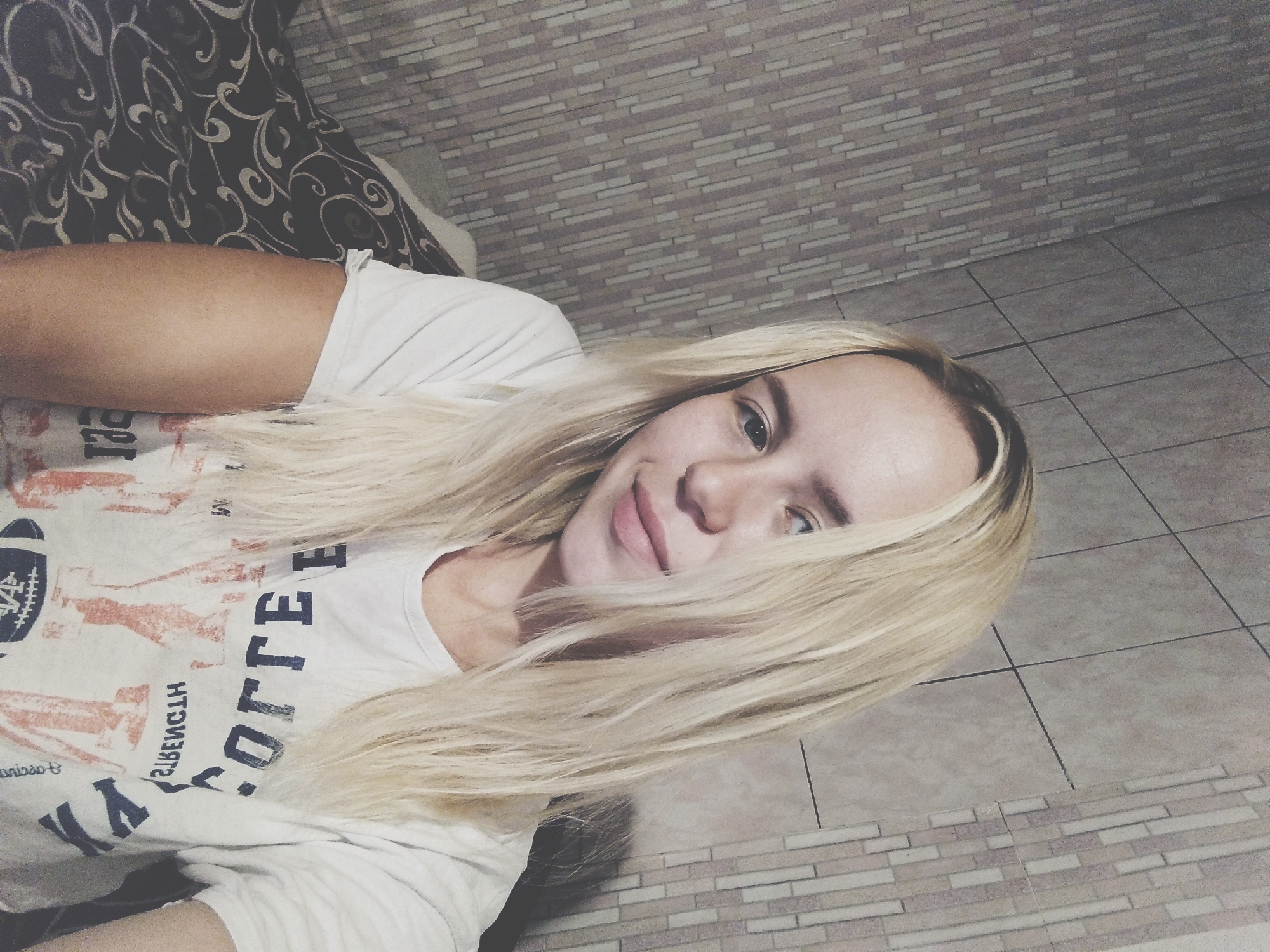A young woman with long, blonde hair is taking a selfie in a room with tiled walls. She is wearing a white t-shirt with a graphic print that includes the word "COLLEGE" and images of footballs and text. Her expression is neutral, and she is looking directly at the camera. The background appears to be a wall with a patterned tile design. The image is dimly lit, and a portion of a patterned blanket or bedding is visible in the lower left corner. The location is specified as Radisne, Ukraine.