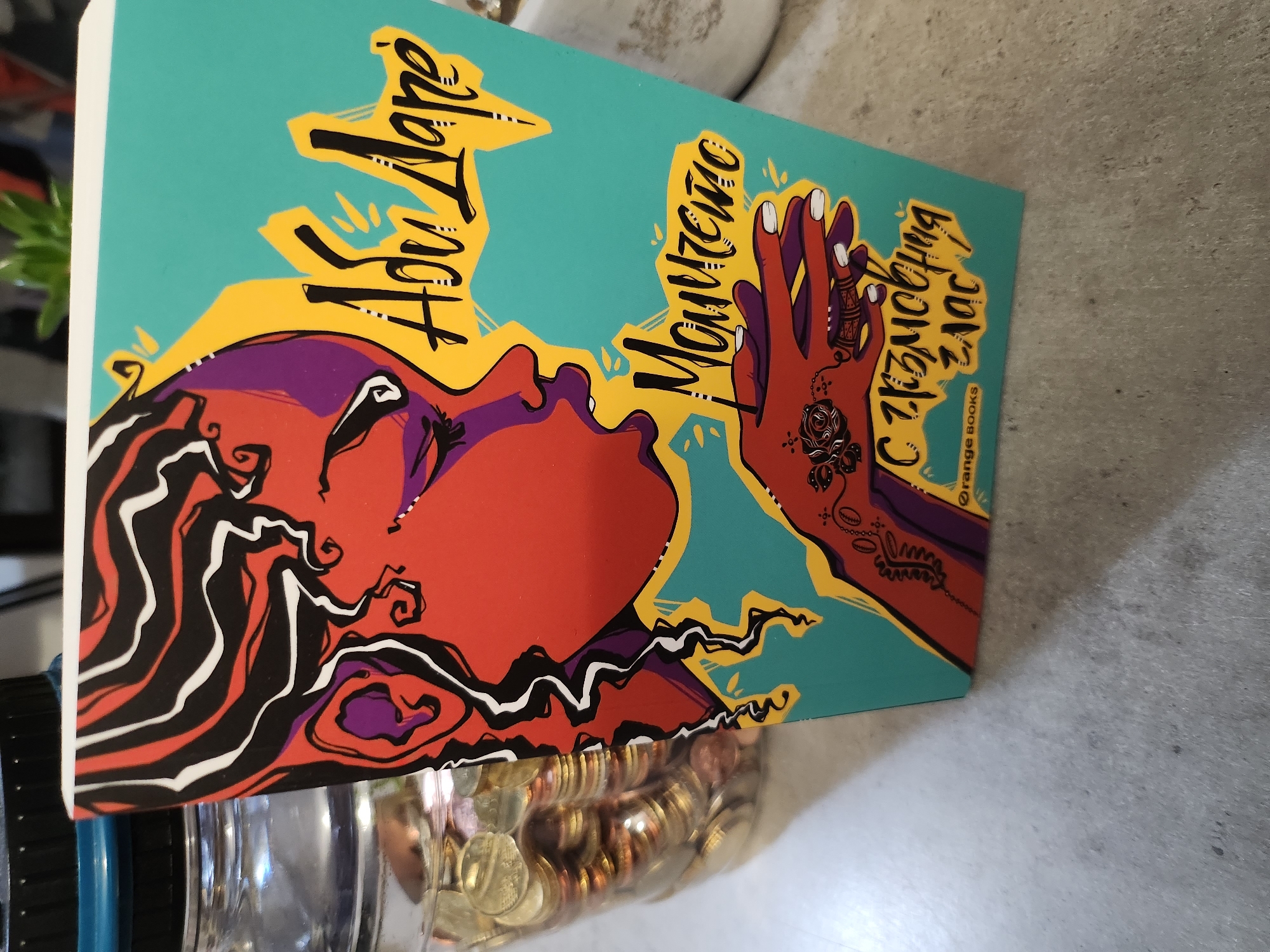 The image shows the front cover of a book tilted slightly to the right, placed on a grey, textured surface, possibly a countertop. To the left of the book, partially visible, is a clear glass jar filled with coins, with a dark blue lid. The book cover features a stylized illustration of a person's face in profile with vibrant colors, including red, purple, black, and white. The text on the cover is in a stylized font and appears to be in Bulgarian. The words "Абу Даре" are prominent in yellow, followed by "Момичета" in a darker, script-like font. Below this, there is text that reads "С гръмогласна глас!". In the bottom left corner of the book cover, a small logo reads "orange books". The lighting suggests an indoor setting with natural light coming from the left. The overall scene is a close-up shot of the book and its immediate surroundings. There is no identifiable location within the city of Tsarevo, Bulgaria, visible in this image.