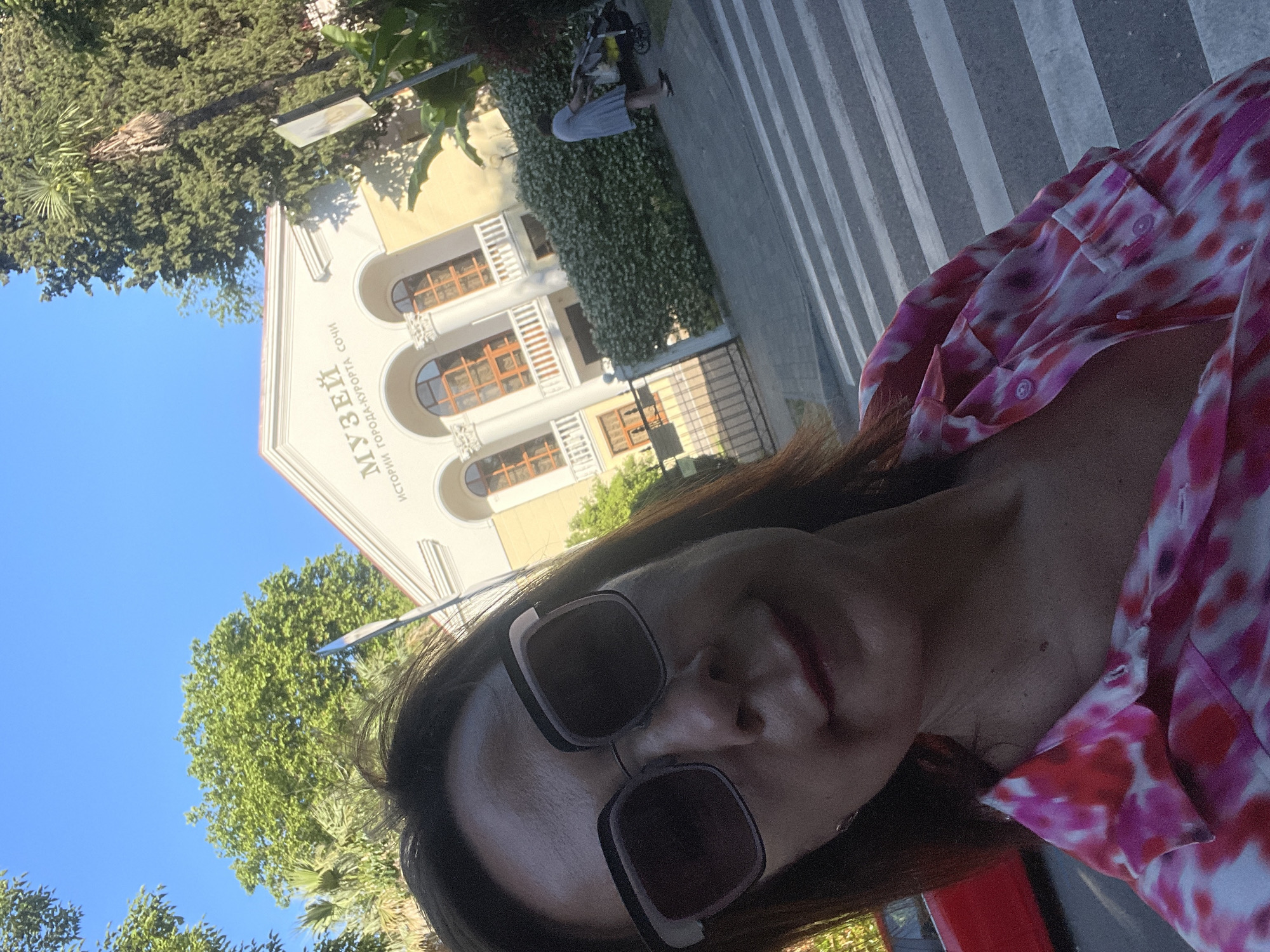 A vertical, low-angle selfie captures a woman in sunglasses and a pink and white tie-dye shirt. She is outdoors, with a bright blue sky above and lush green trees framing the left side of the image. Behind her is a light-yellow building with arched windows and columns, identified by text on its facade. The text, which appears to be in Russian, reads "МУЗЕЙ ИСТОРИИ ГОРОДА-КУРОРТА СОЧИ", translating to "MUSEUM OF THE HISTORY OF THE RESORT CITY OF SOCHI". The setting appears to be sunny, suggesting daytime. The woman's face is the primary focus, with her sunglasses obscuring her eyes. Her expression is neutral, and the overall mood is casual and relaxed. In the background, a paved area with a striped pattern and a person with a stroller are visible, indicating a public space, possibly a promenade or a park area near the museum.