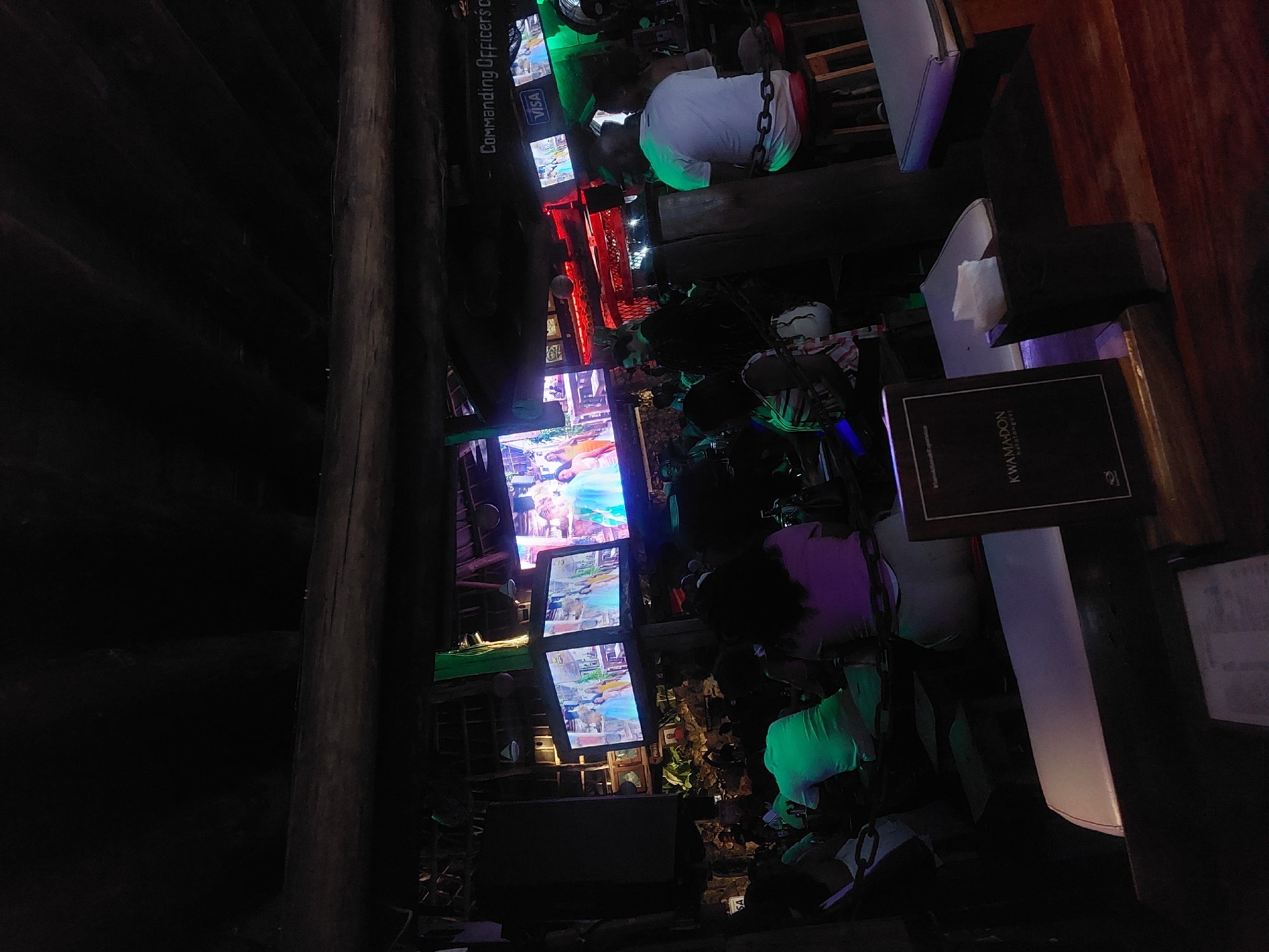 The image depicts a bar scene with patrons in Dar es-Salaam, Tanzania. Multiple screens display content, including the VISA logo. The environment suggests a lively, possibly tourist-oriented establishment. Patrons are seated and casually dressed. A hanging seat adds a distinct feature to the scene. A menu with "KWAMADON" is visible. The bar is dimly lit, and the structure is a combination of wood and modern elements.