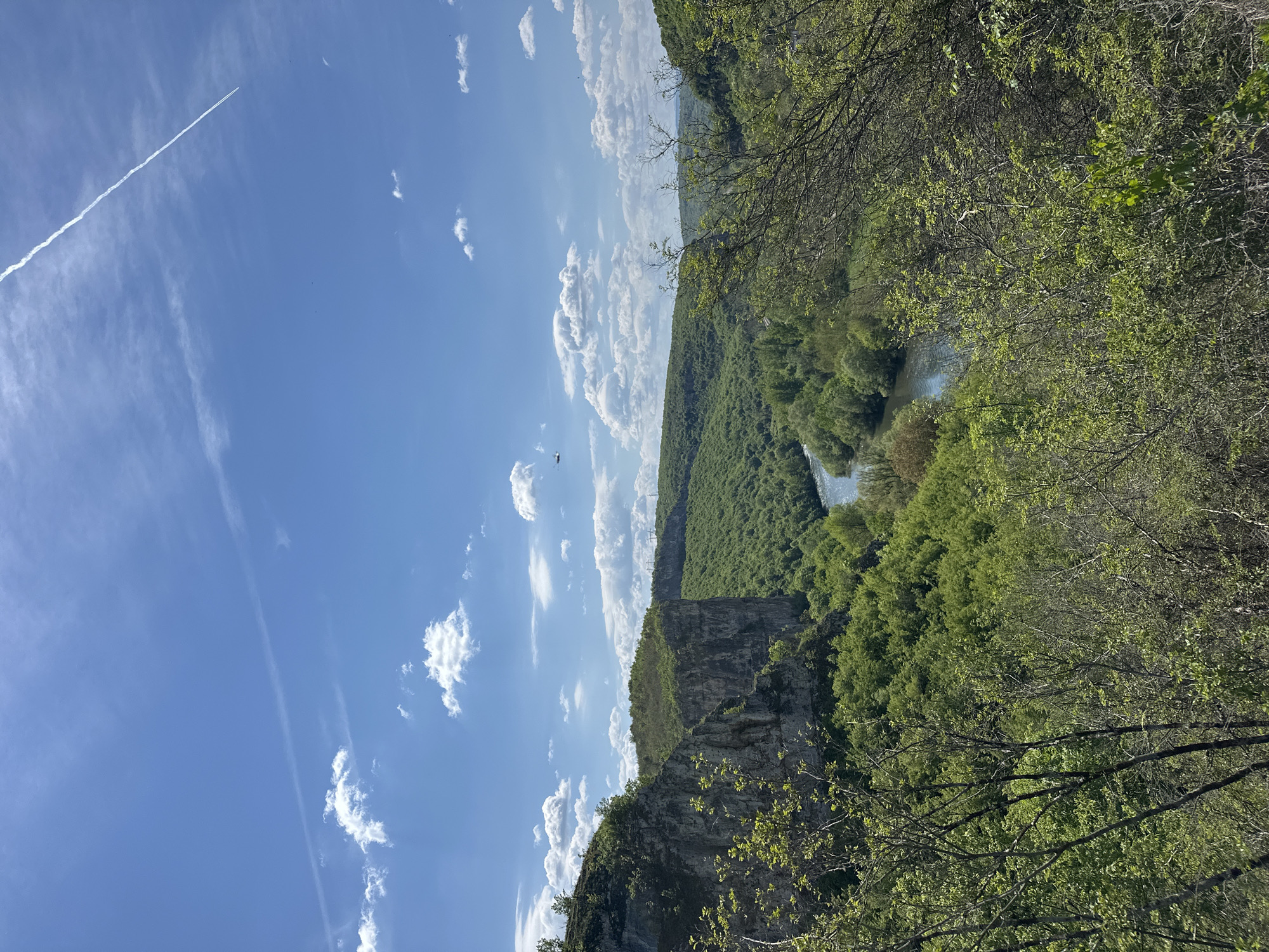 The image depicts a scenic landscape in Karlukovo, Bulgaria. A river meanders through a canyon, with steep cliffs and dense green forests lining both sides. The sky is bright blue with scattered white clouds and a contrail from an aircraft. A small helicopter is visible in the distance. The perspective is from an elevated position.