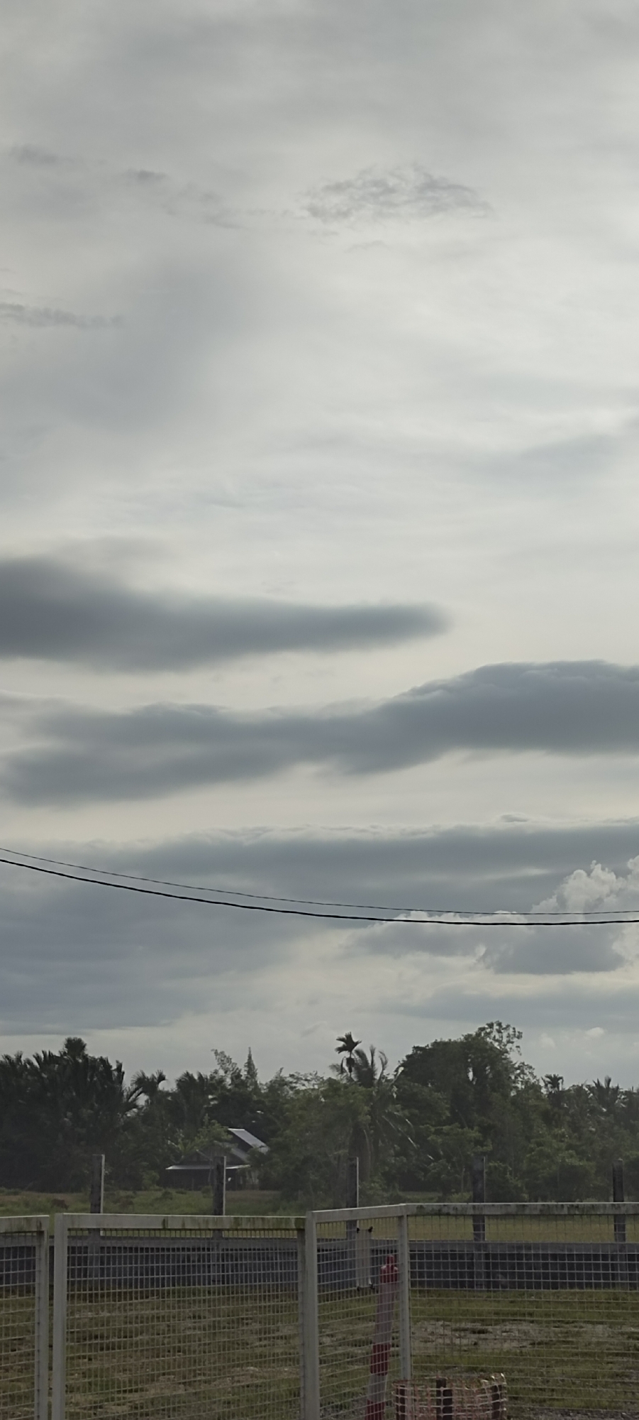 A muted, cloudy day in Meunasahaluedrienlhoksukon Barat, Indonesia, is captured in this outdoor shot. A wire fence, prominently in the foreground, is composed of gray, rectangular mesh panels supported by white vertical posts. A patch of grassy terrain lies behind the fence. Behind the fence, there is an area covered with a variety of trees and foliage. Partially visible is a structure, possibly a house or building, amidst the vegetation. Above the treeline, a gray, overcast sky is evident, with layered clouds creating a textured backdrop. Power lines stretch across the scene between the trees and the clouds.