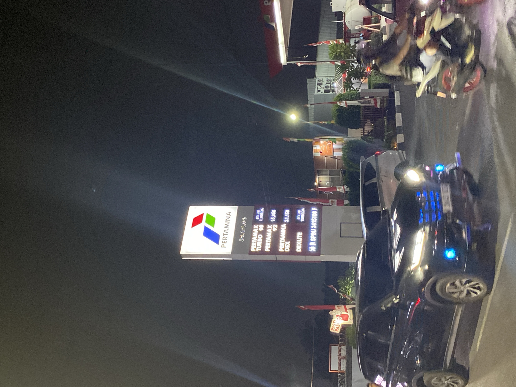The image shows a Pertamina gas station in Tangerang, Indonesia, at night. A tall sign displays fuel prices. Pertamax Turbo 98 is listed as "BELUM TERSEDIA" (not available). Pertamax 92 costs 16,950, Pertamina Dex costs 21,150, and Dexlite is also "BELUM TERSEDIA". A black SUV is parked in the foreground, its headlights illuminated with blue accents. To the right, a person rides a motorcycle. The background shows more gas station structures, flags, and streetlights. The overall scene is characteristic of a typical urban gas station at night.