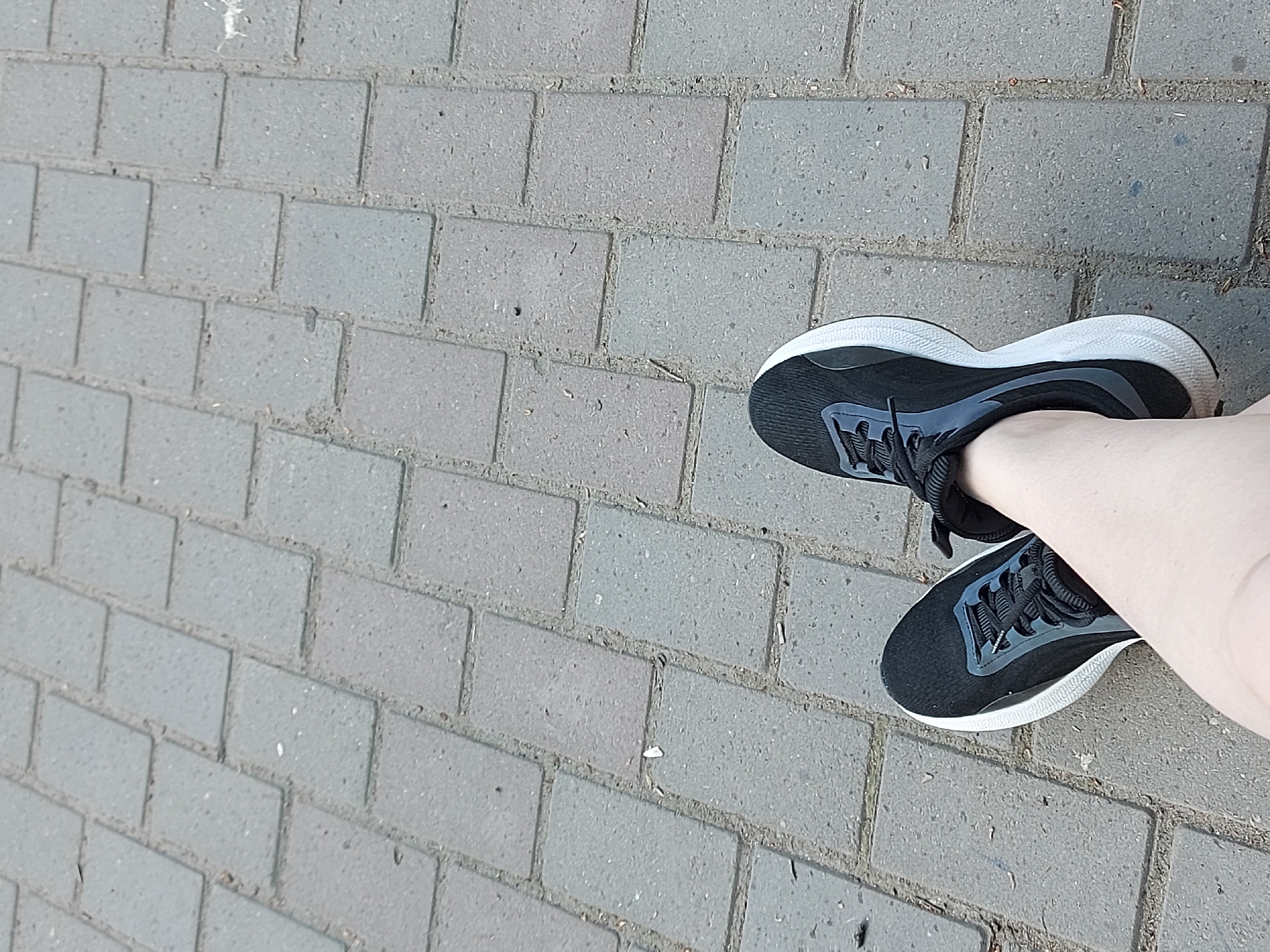 The image shows a pair of legs wearing black and grey athletic shoes standing on a paved surface made of grey rectangular bricks. The bricks are arranged in a running bond pattern. The shoes have white soles and black laces. The legs are pale. The location is suggested to be Chaikovsky, Russia, based on the prompt, but no features in the image explicitly confirm this location. There are no other people, subjects, objects, actions, or interactions visible in the image. The environment is limited to the paved surface.