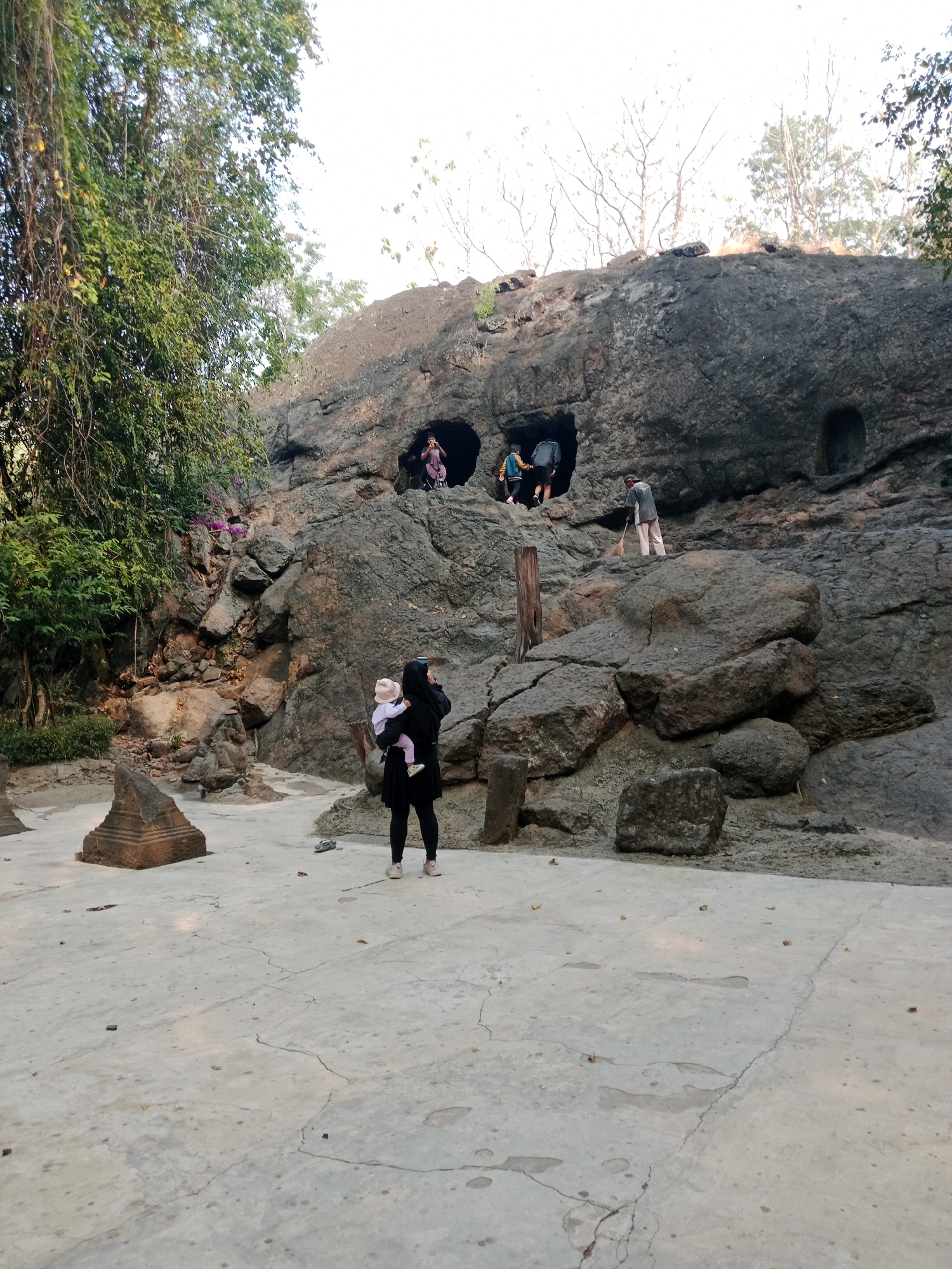 The image depicts a large, rough rock face with several openings, resembling caves or alcoves, carved into it. At the base of the rock face, a woman in black clothing and a headscarf is holding a baby. She is facing away from the camera, looking towards the rock face and holding a phone up, seemingly taking a picture or video. To her left, a small, dark, pyramid-shaped stone structure is visible on the concrete ground. In front of the rock face, two children in casual clothing are entering one of the openings, with one appearing to be climbing. Further to the right and slightly higher up on the rock face, a person is seen sweeping with a broom. The environment appears to be an outdoor, somewhat rustic location, possibly a park or historical site, with trees and foliage visible in the background. The ground is paved with concrete, which shows cracks and some scattered leaves. The overall lighting suggests it is daytime. The location is identified as Gua Selomangleng in Kediri City, Indonesia.