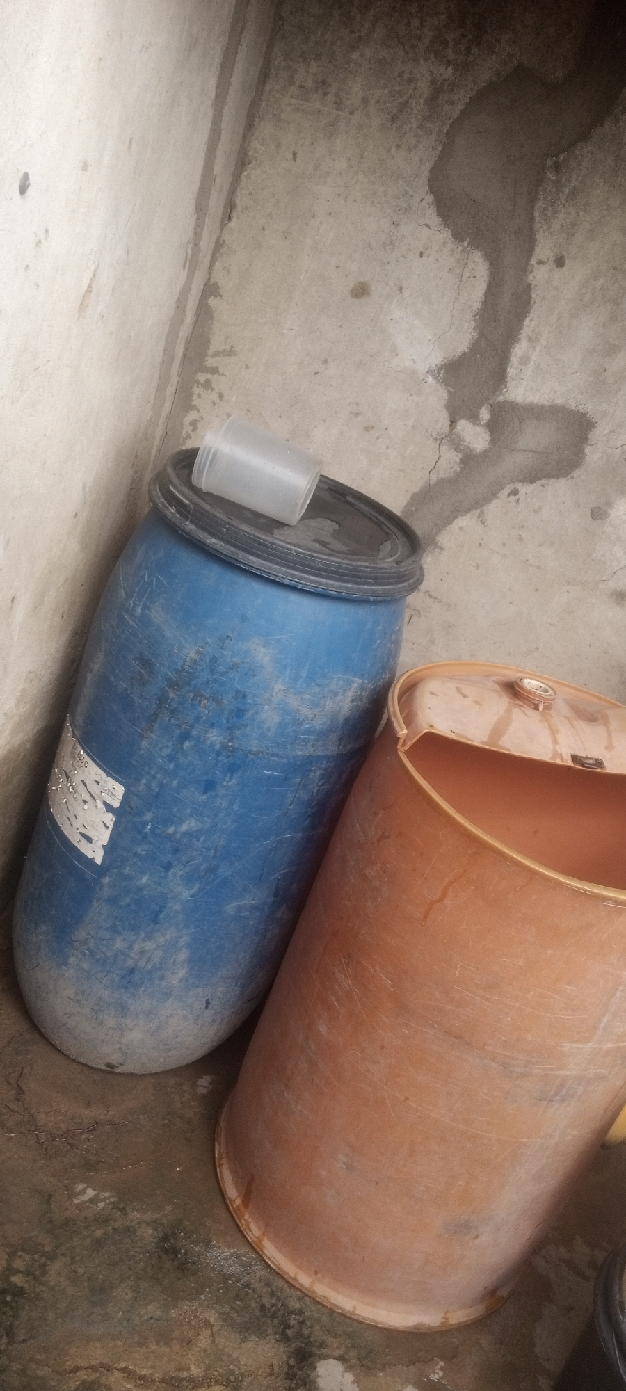 The scene depicts two plastic barrels in what appears to be a corner, likely in a storage area. One barrel is blue with a black lid and a small translucent container placed on top. A partially damaged label is affixed to its side. The other barrel is orange and appears to be open. The barrels are situated against what looks like a concrete wall, which has visible cracks and repairs. The floor is also concrete and appears damp. The location is specified as Sabon Tasha, Nigeria, but there are no elements visible in the image that directly confirm this, and there are no people, interactions, or actions visible.