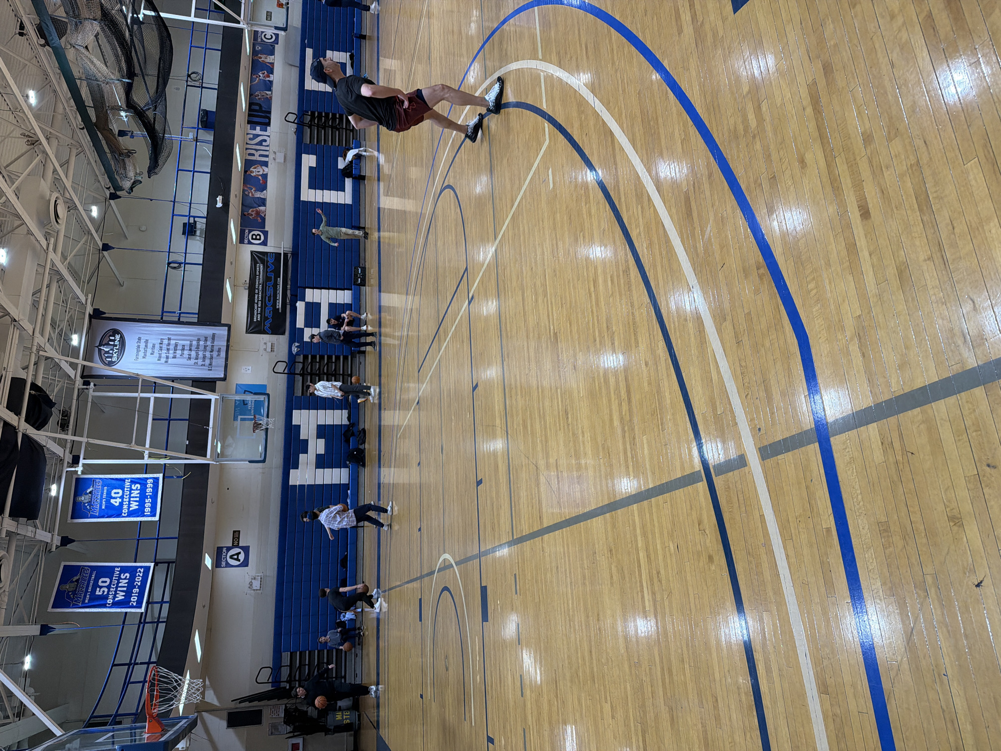 The image captures the interior of a gymnasium, possibly located in the city of New York, United States of America, as indicated by the "SKYLINE" banner listing New York locations. The gymnasium has a wooden basketball court with blue and white court markings. There are several people present, some standing and others sitting on blue bleachers along the side of the court. A man is visible in the upper part of the frame walking on the court. Banners and signs are displayed on the wall, including ones that celebrate achievements of "MACCABEES" in men's tennis, such as "40 Consecutive Wins 1995-1999" and "50 Consecutive Wins 2019-2022." The environment includes basketball hoops with nets, overhead lights, and ventilation systems visible on the ceiling. The general action is a warm-up or practice setting for a basketball or tennis team.